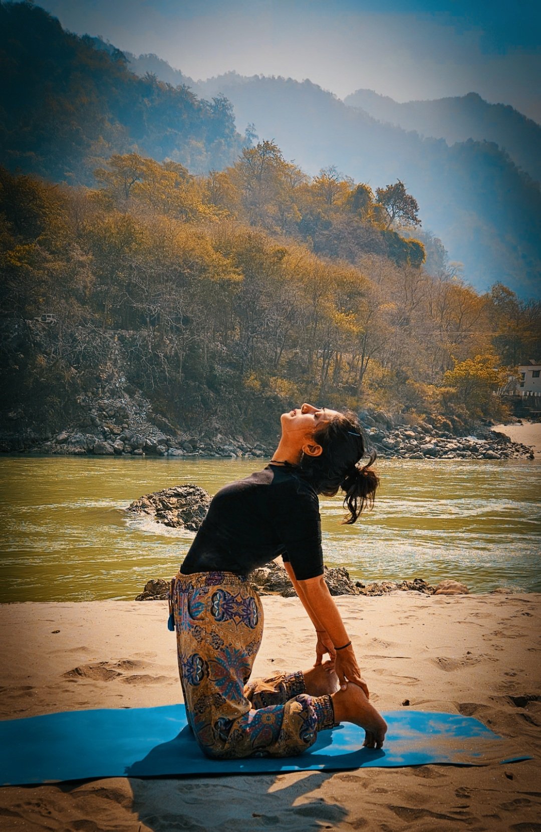 A woman practicing yoga on a beach by a river, with mountains and trees in the background during sunset or early morning.