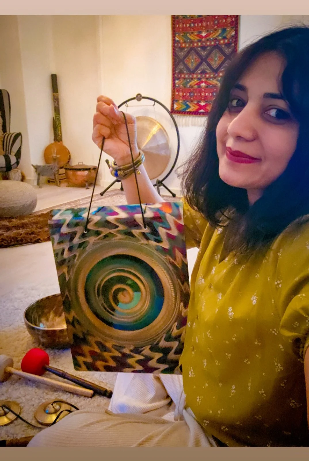 A woman with dark hair and red lipstick smiling while holding a colorful, abstract-patterned wind chime in a living room. In the background, there is a gong, a large wooden stringed instrument, a tapestry on the wall, and various musical instruments on the floor.