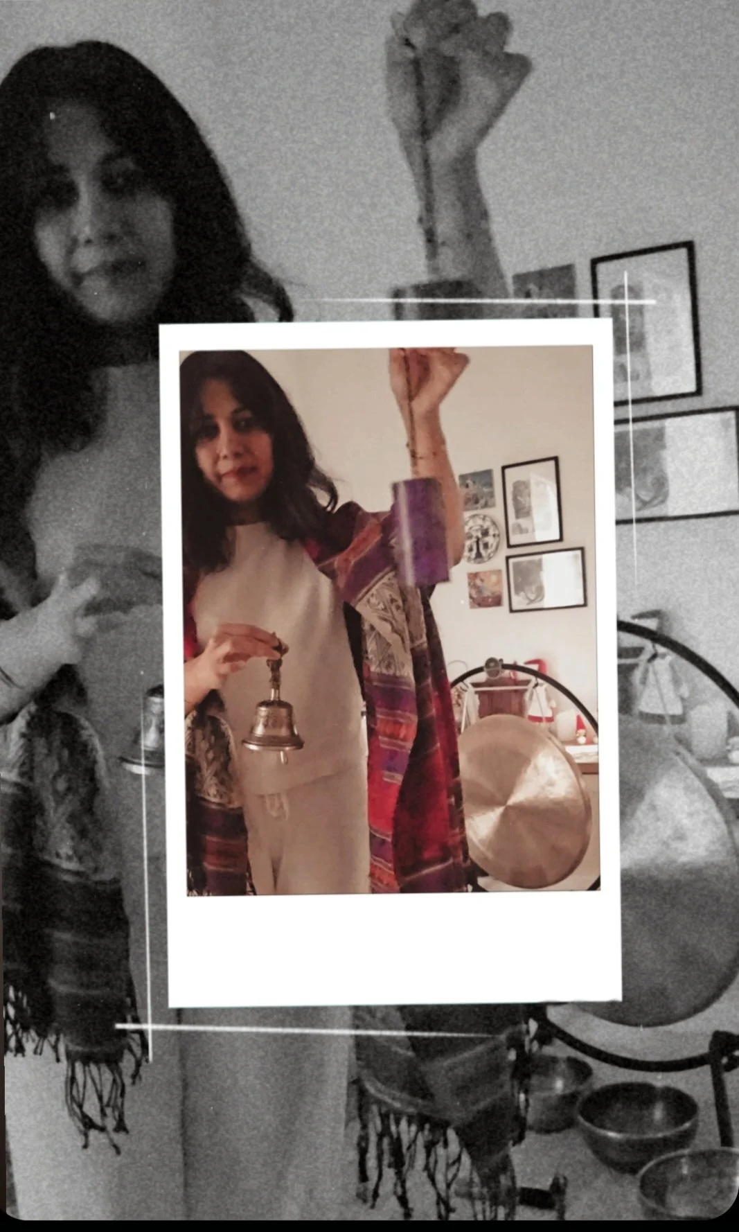 A woman holding a small bell in front of a colorful wall with framed artwork, standing in a room with a gong and drums in the background.
