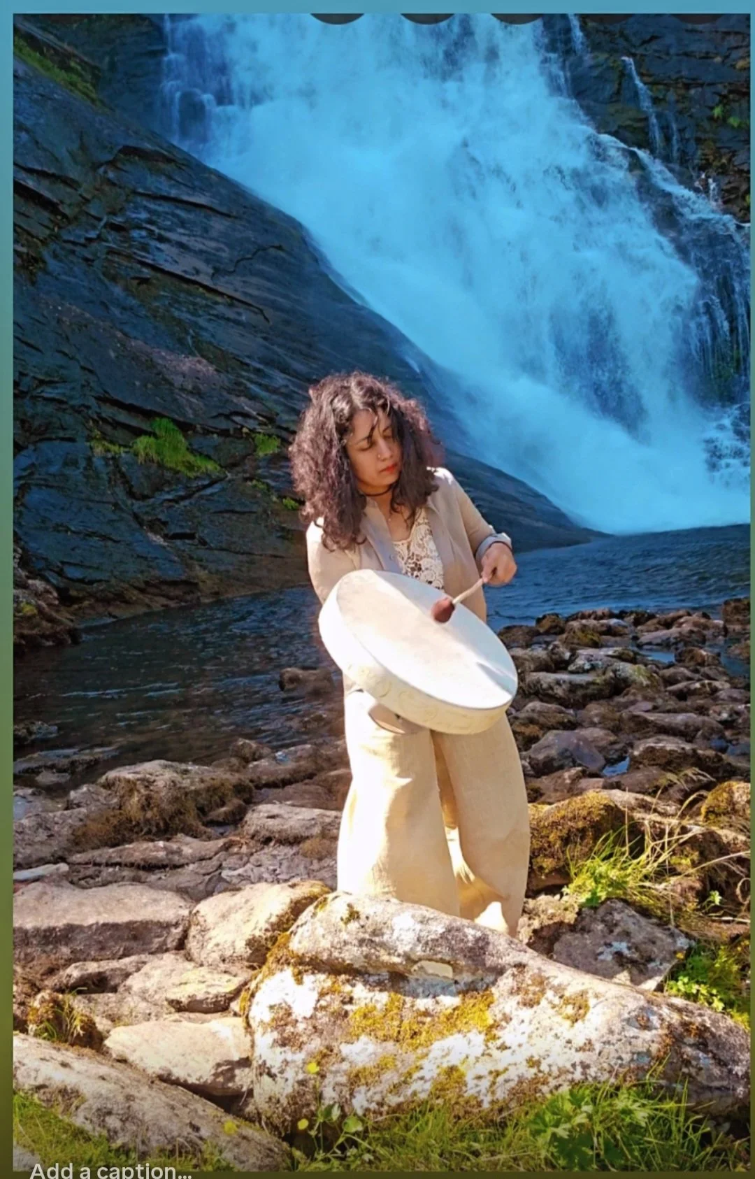 A woman playing a drum by a waterfall in a rocky, natural setting.