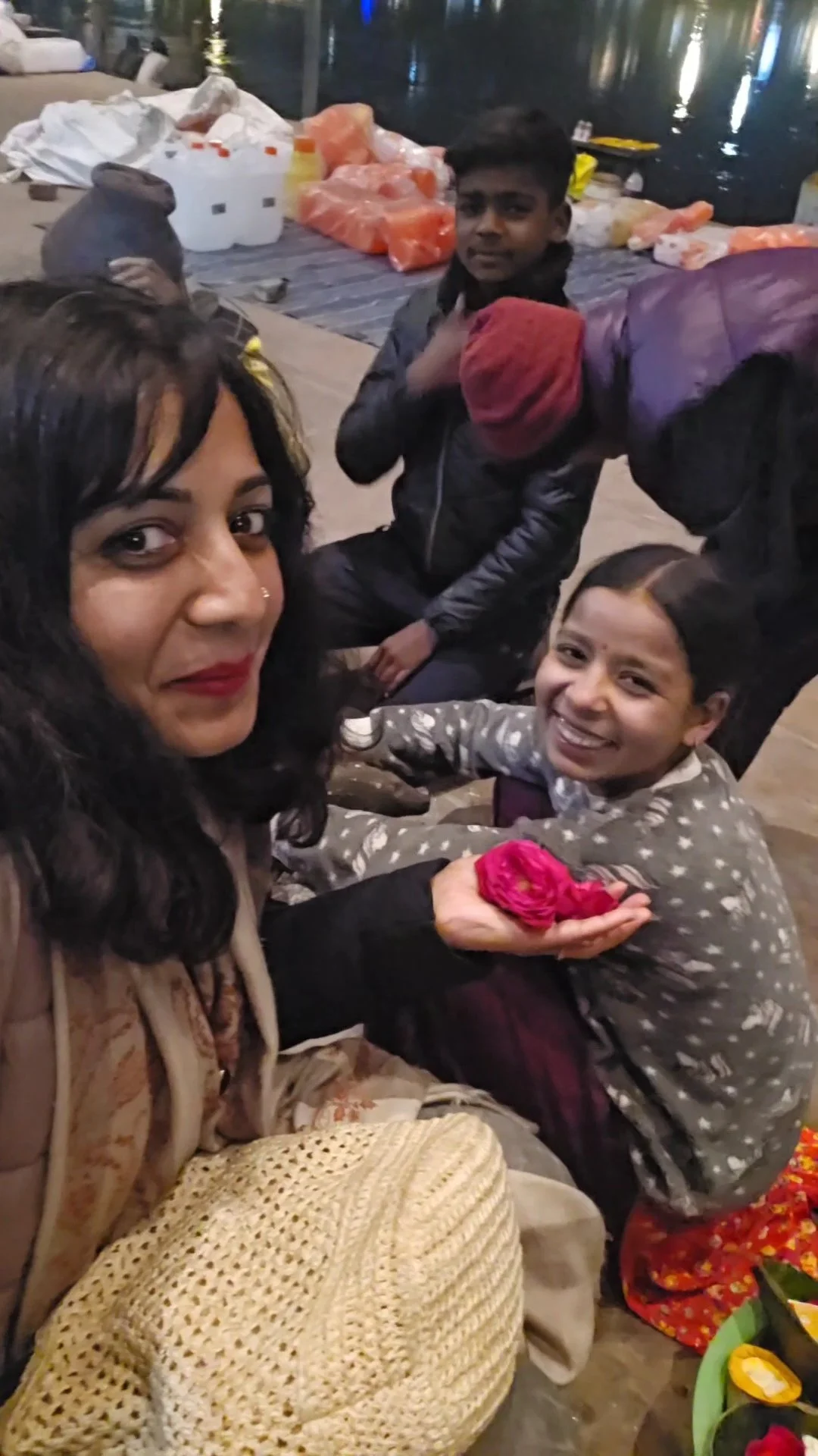 A woman and two smiling children at a gathering near water at night, with supplies and items in the background.