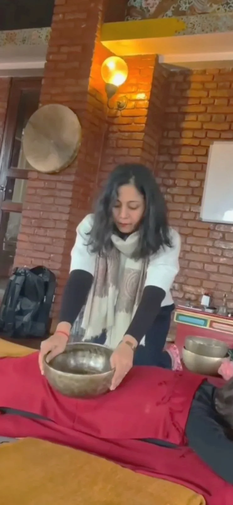 A woman with shoulder-length dark hair, wearing a beige scarf and a black and white long-sleeve shirt, is holding a metal singing bowl with both hands. She is standing indoors with a brick wall and warm lighting, and there are other bowls and objects on the table in front of her.