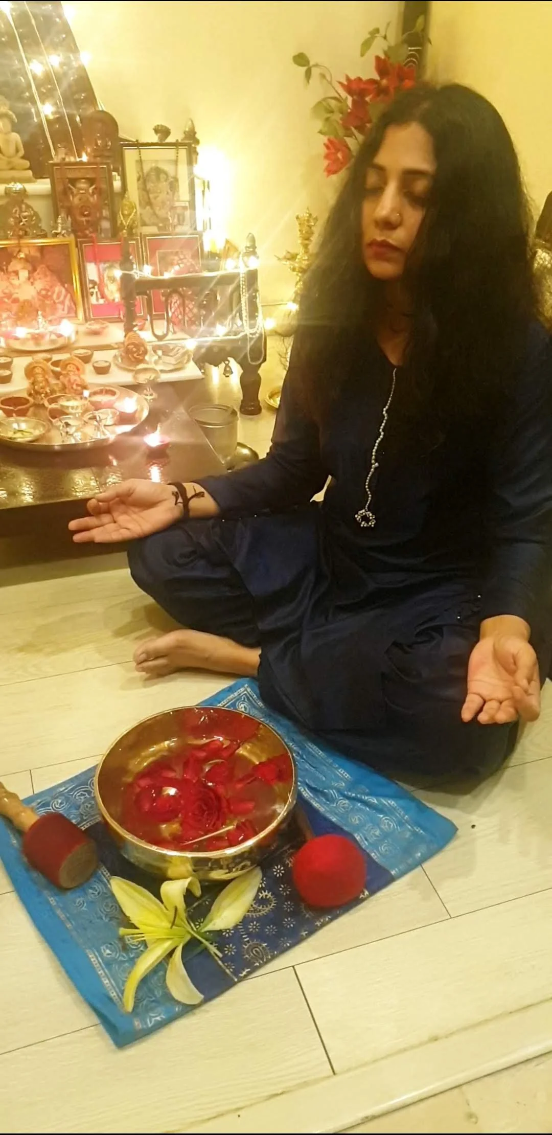 A woman seated cross-legged on the floor in front of a spiritual altar, with candles, statues, and framed images, meditating with closed eyes. In front of her is a bowl with red flower petals, and decorative items including yellow lilies and red balls. The scene is illuminated with warm lighting.