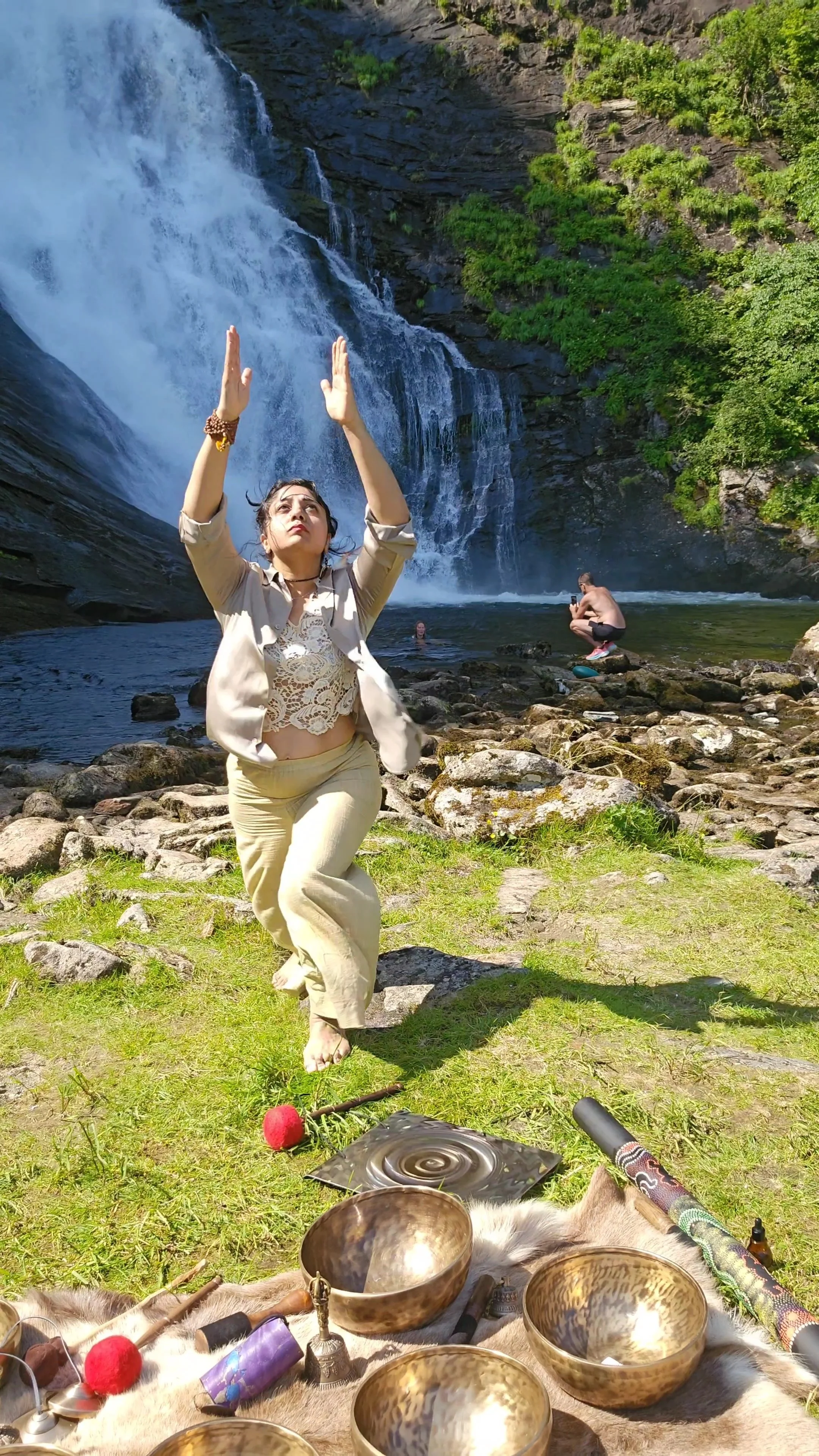 A woman practicing yoga or meditation in front of a waterfall, with other people swimming in the background, and musical instruments and ritual objects on the ground.