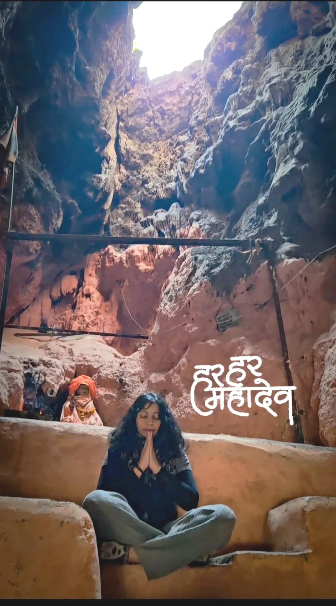 A woman practicing meditation inside a cave with red and brown rocks. She is sitting cross-legged with hands in prayer position, eyes closed. There is some written text in a South Asian script on the right side of the image.