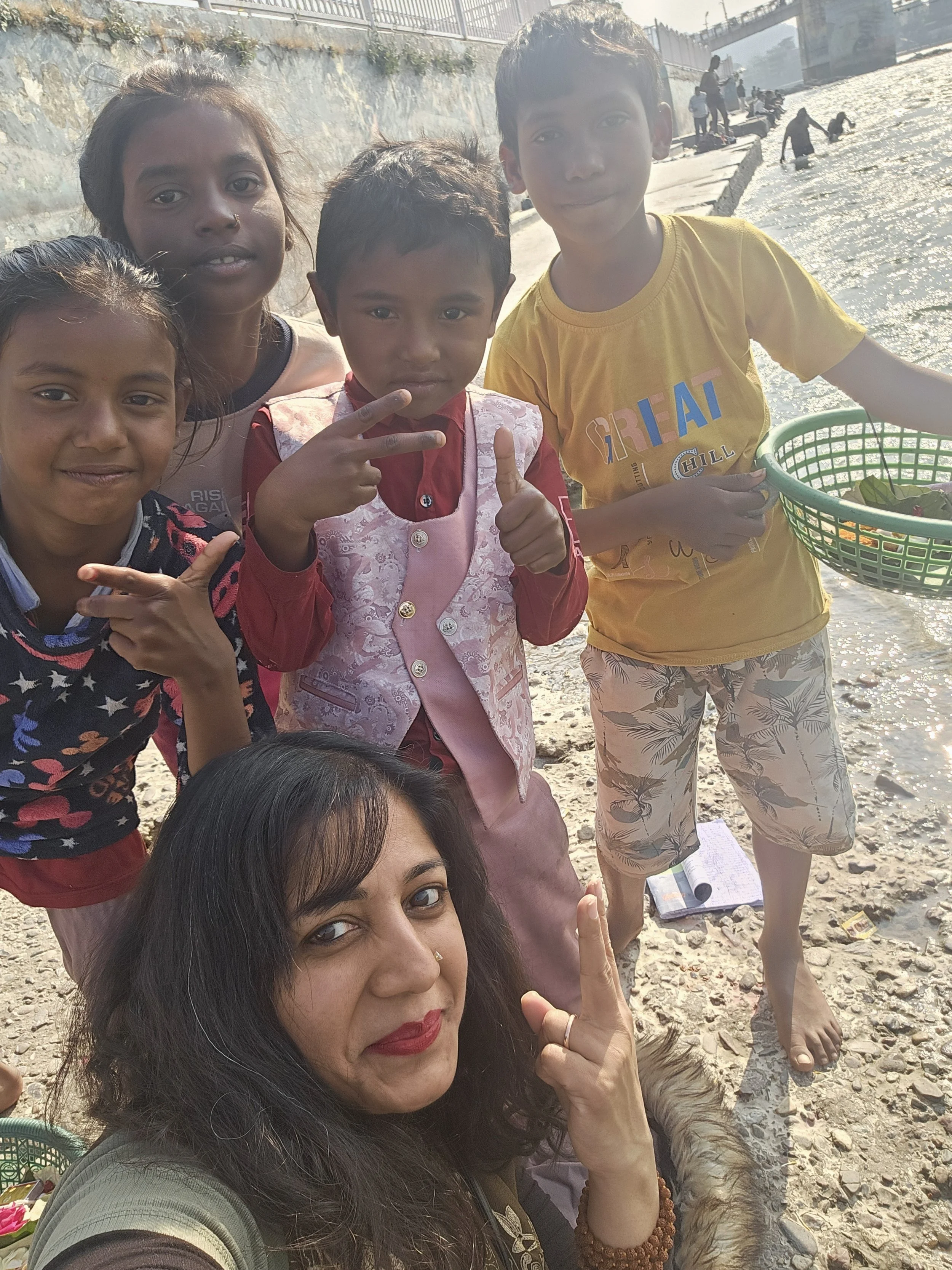 A woman with black hair and red lipstick taking a selfie with four children on a beach. The children are smiling and making peace signs, with the sea and other people in the background.