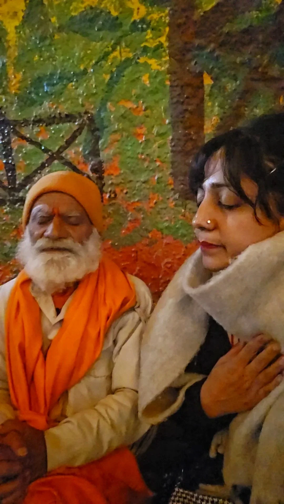 A woman with dark curly hair, wearing a beige scarf, sitting with her eyes closed, and a bearded old man with a yellow turban, orange scarf, and traditional clothing, seated outdoors at night with autumn leaves in the background.