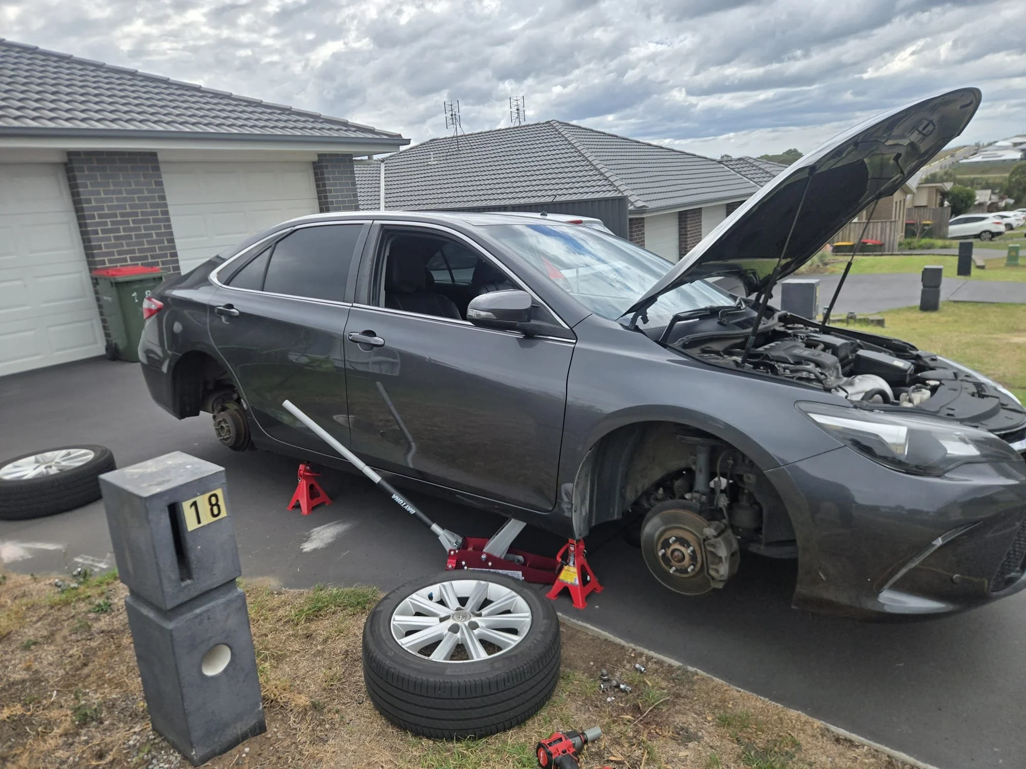 A dark gray sedan with its front wheels removed and placed on the ground, supported by red jack stands, is in a driveway in front of a house with a garage door. The car's hood is open, revealing the engine, and the front right wheel is missing. There are two used tires on the ground and tools, including a power drill, nearby. A concrete post in the foreground has a yellow and black label with the number 18. The background shows a cloudy sky over a suburban neighborhood with other houses and parked cars.