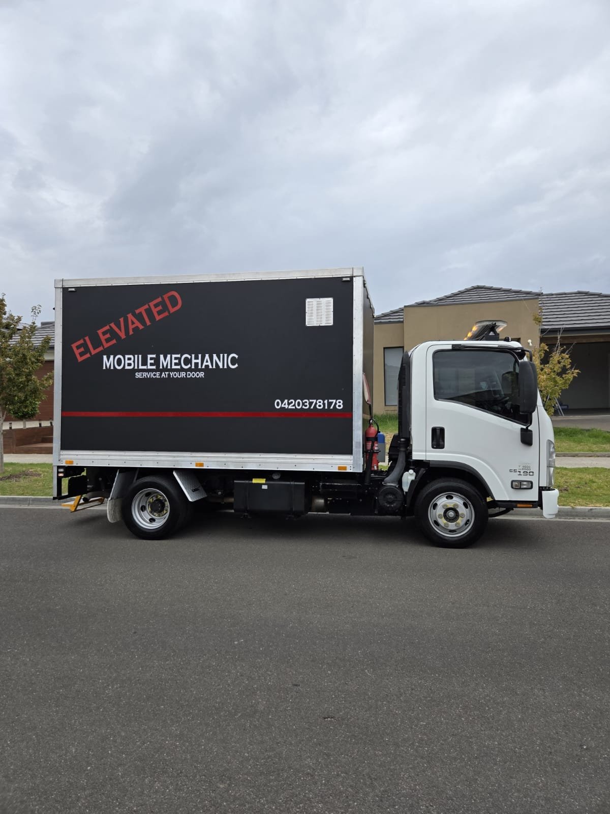 A mobile mechanic service truck parked on the street in front of residential houses. The truck is white with a large black and red sign on the side that reads 'ELEVATED MOBILE MECHANIC' and includes a phone number.