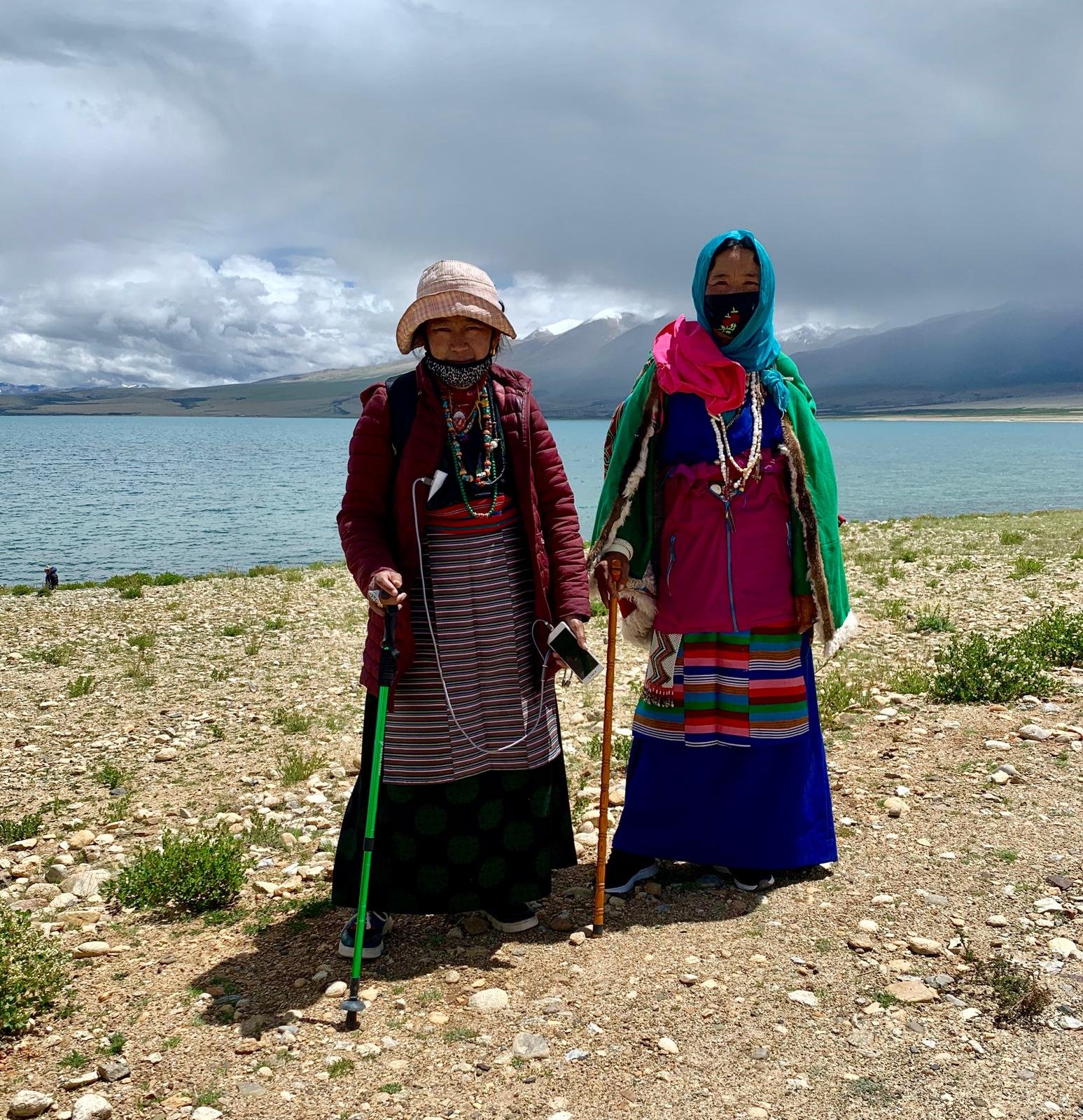 Two elderly women in traditional colorful clothing standing by lake Monasarovar with mountains and cloudy sky in the background.