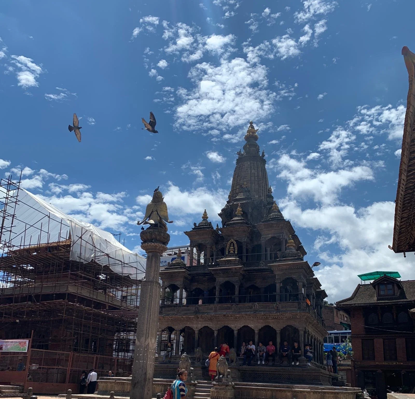 A historic temple with ornate carvings and a tall central spire, set against a blue sky with scattered clouds. There are people seated on the steps and walking around the temple, with scaffolding on the left side indicating construction or renovation work.