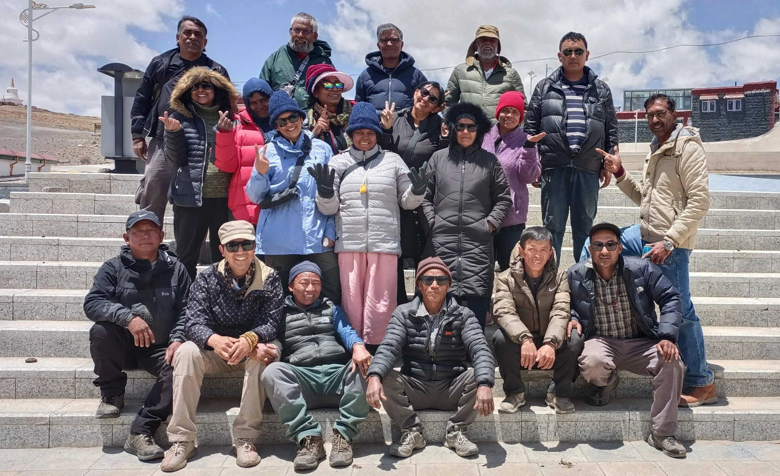 A large group of diverse people posing on outdoor steps at a scenic, mountainous location, wearing outdoor clothing and sunglasses, with a cloudy sky above.