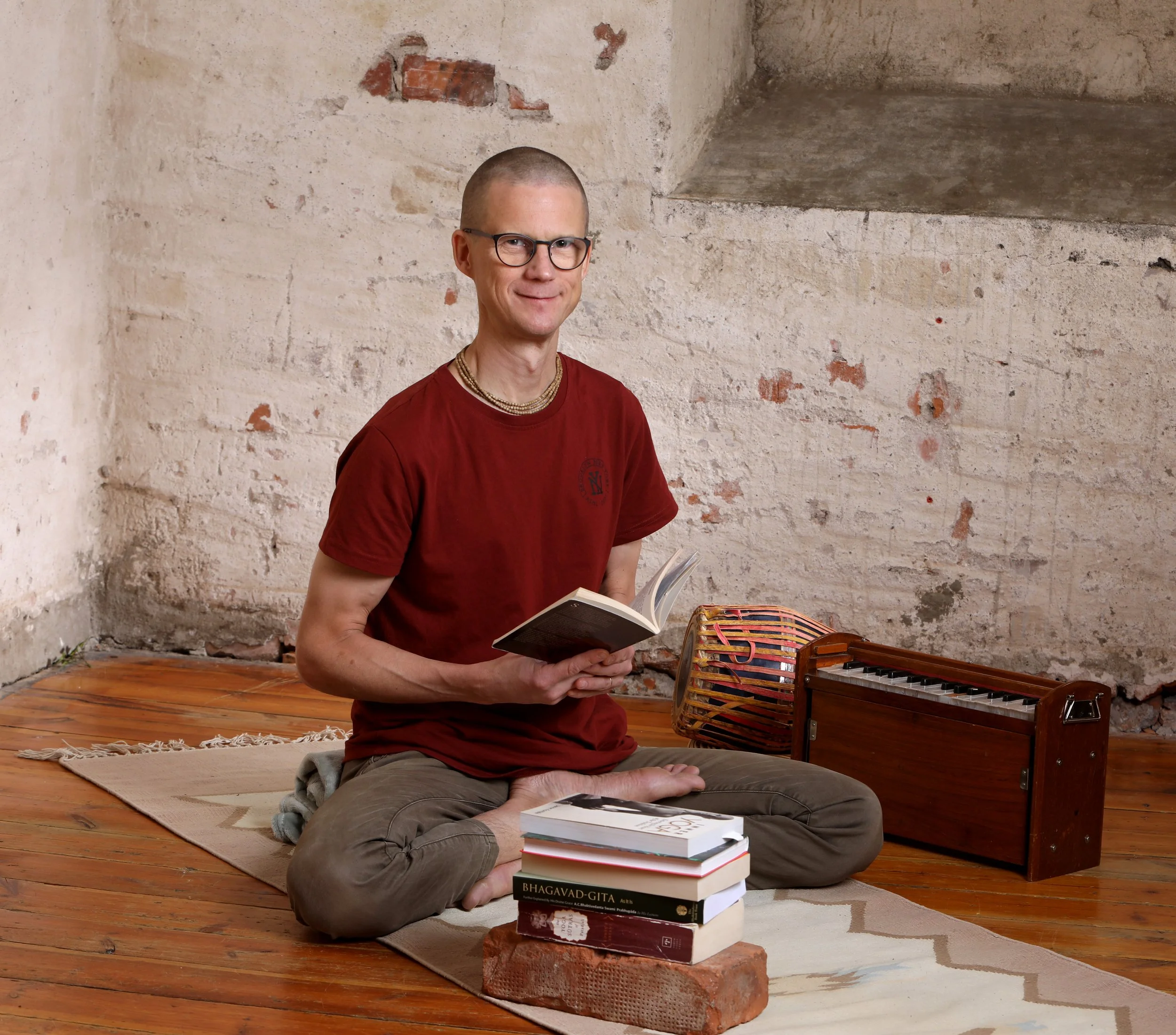 A man sitting on a small rug, holding a book, with a stack of books in front of him. He is in a room with exposed brick and plaster walls, on a wooden floor, with a keyboard on his right.