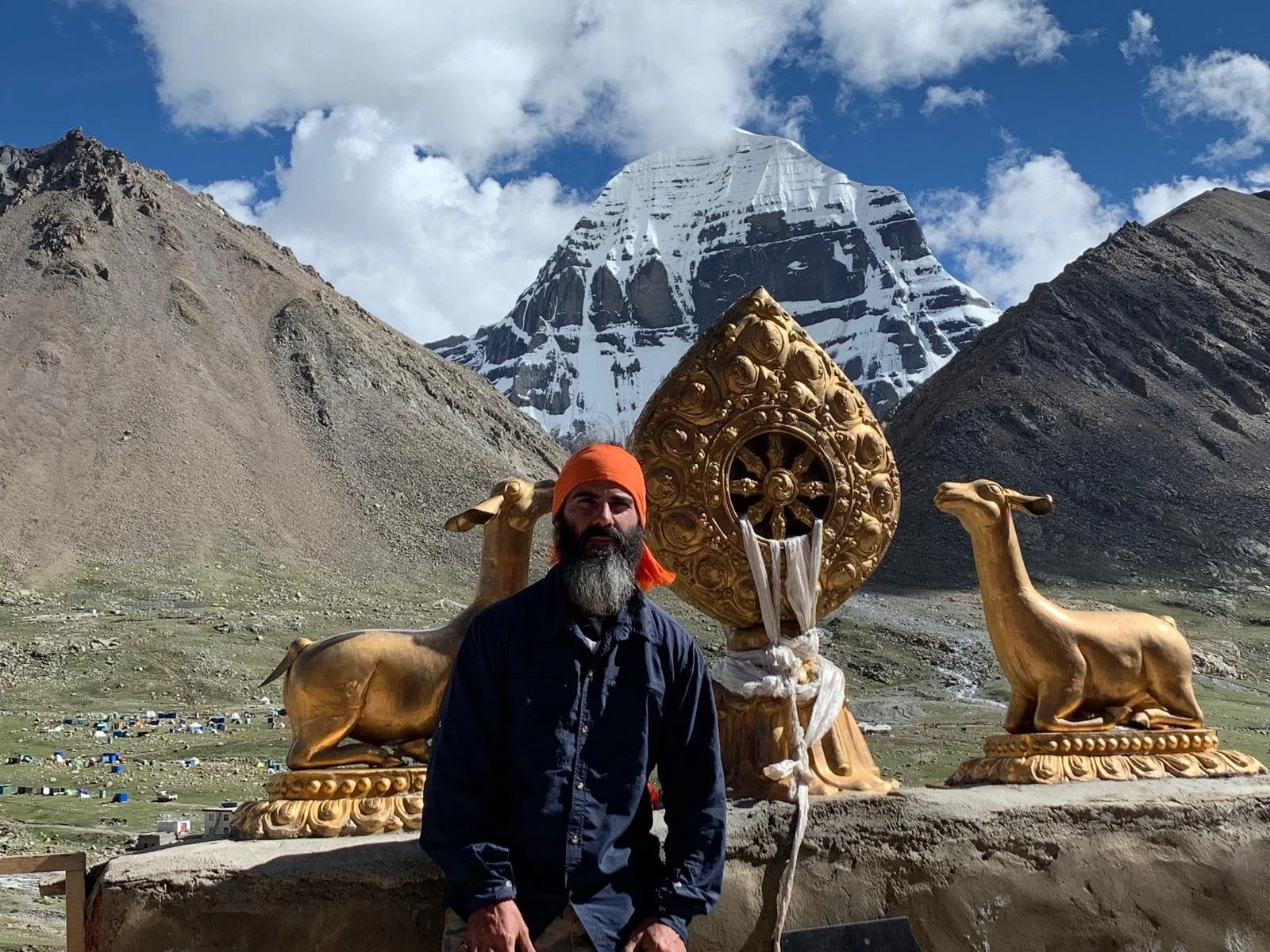 Michael standing in front of large golden religious sculptures of lotus flowers and a wheel with a Mount Kailash in the background.