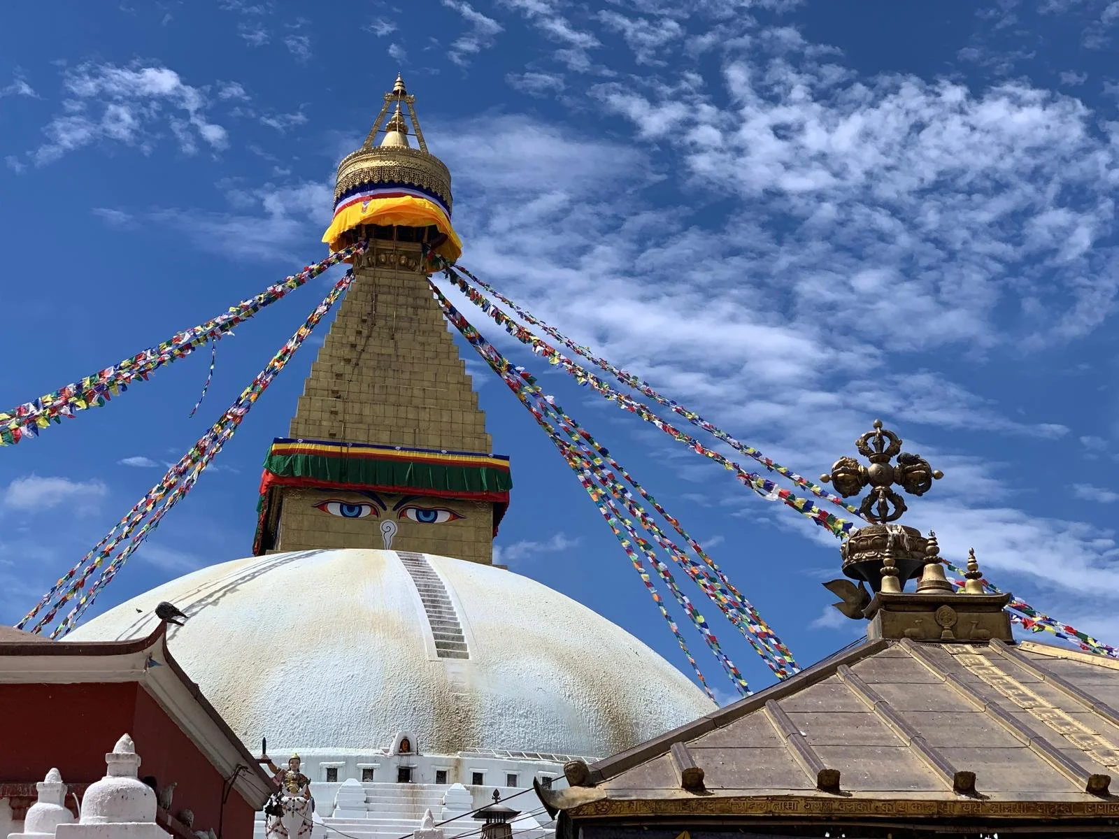 A Buddhist stupa with a golden spire, colorful prayer flags, and an eye symbol on the structure under a blue sky with clouds.