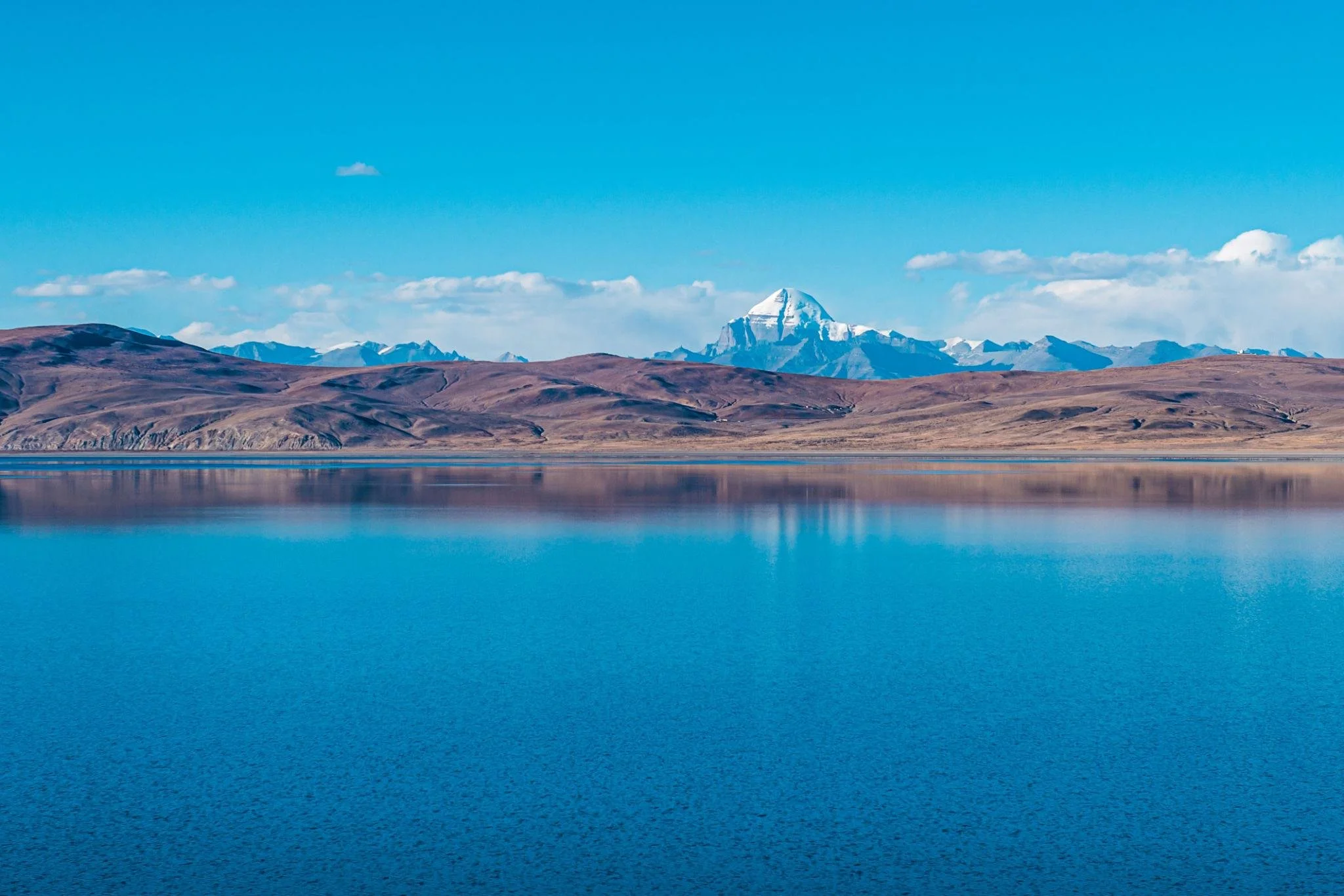 A scenic lake Monasarovar view with calm blue water in the foreground, dry rolling hills in the middle distance, and snow-capped mountains under a bright blue sky with some clouds in the background.