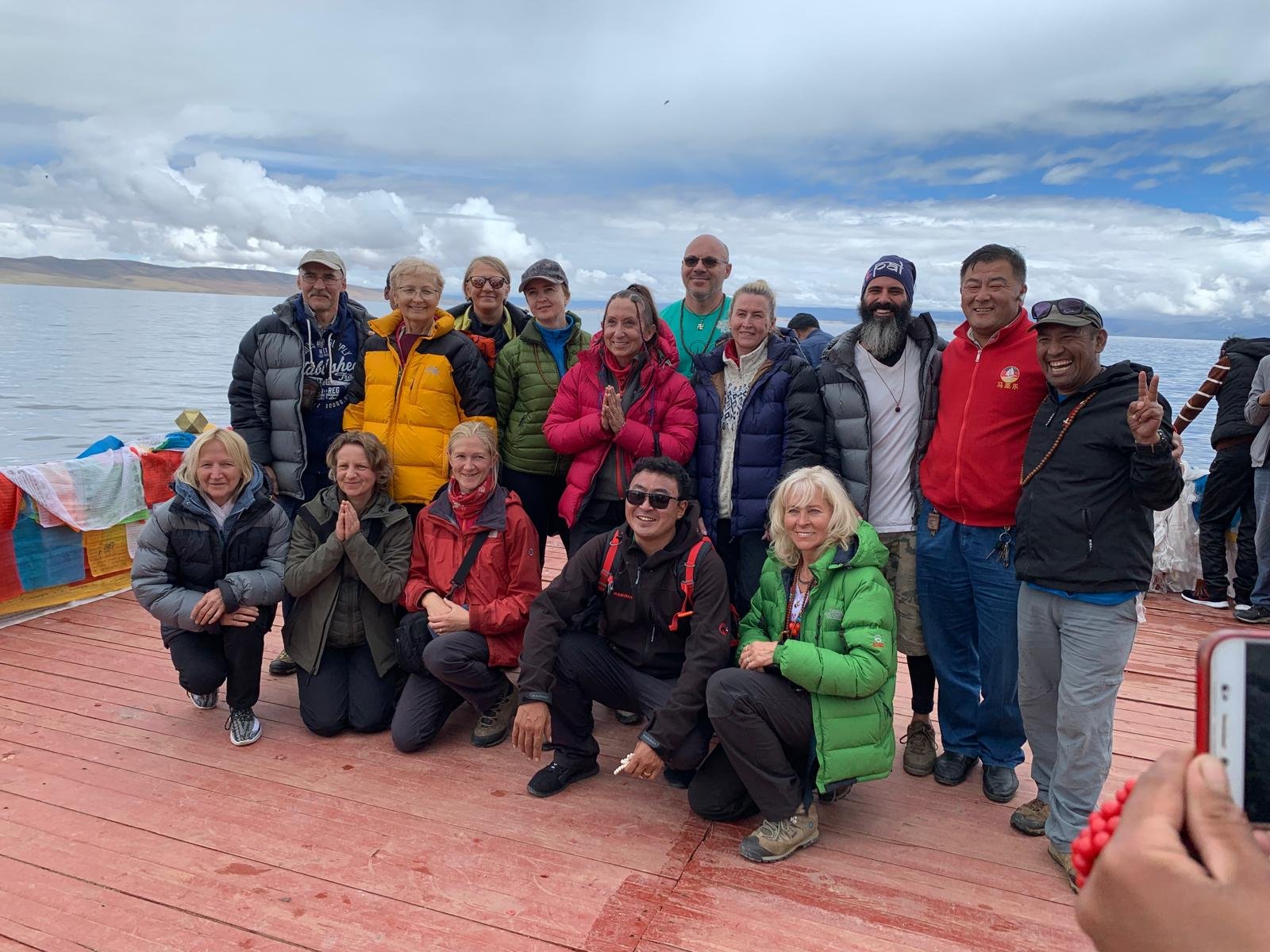 A group of people wearing outdoor jackets poses on a wooden deck by a body of water, with mountains and clouds in the background.