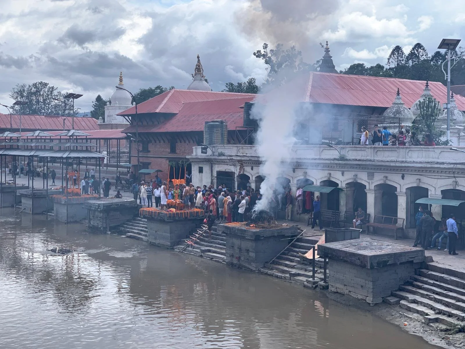 People gathered on the steps of a riverside temple in Nepal, performing rituals and offering prayers during a religious ceremony with smoke and fire offerings. Traditional architecture with temple spires and modern solar streetlights are visible against a cloudy sky.