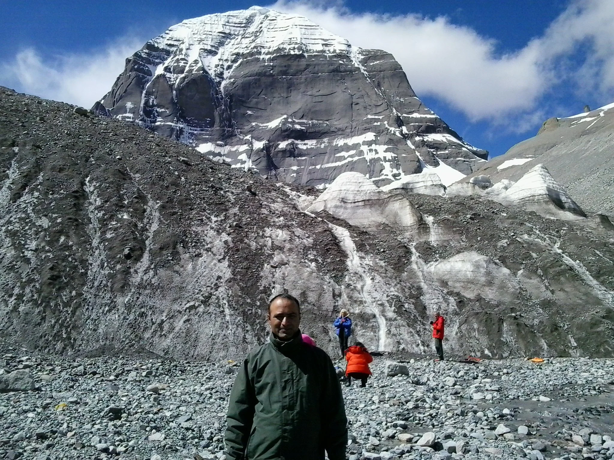 People hiking in a rocky mountain landscape with a Mount Kailash in the background.