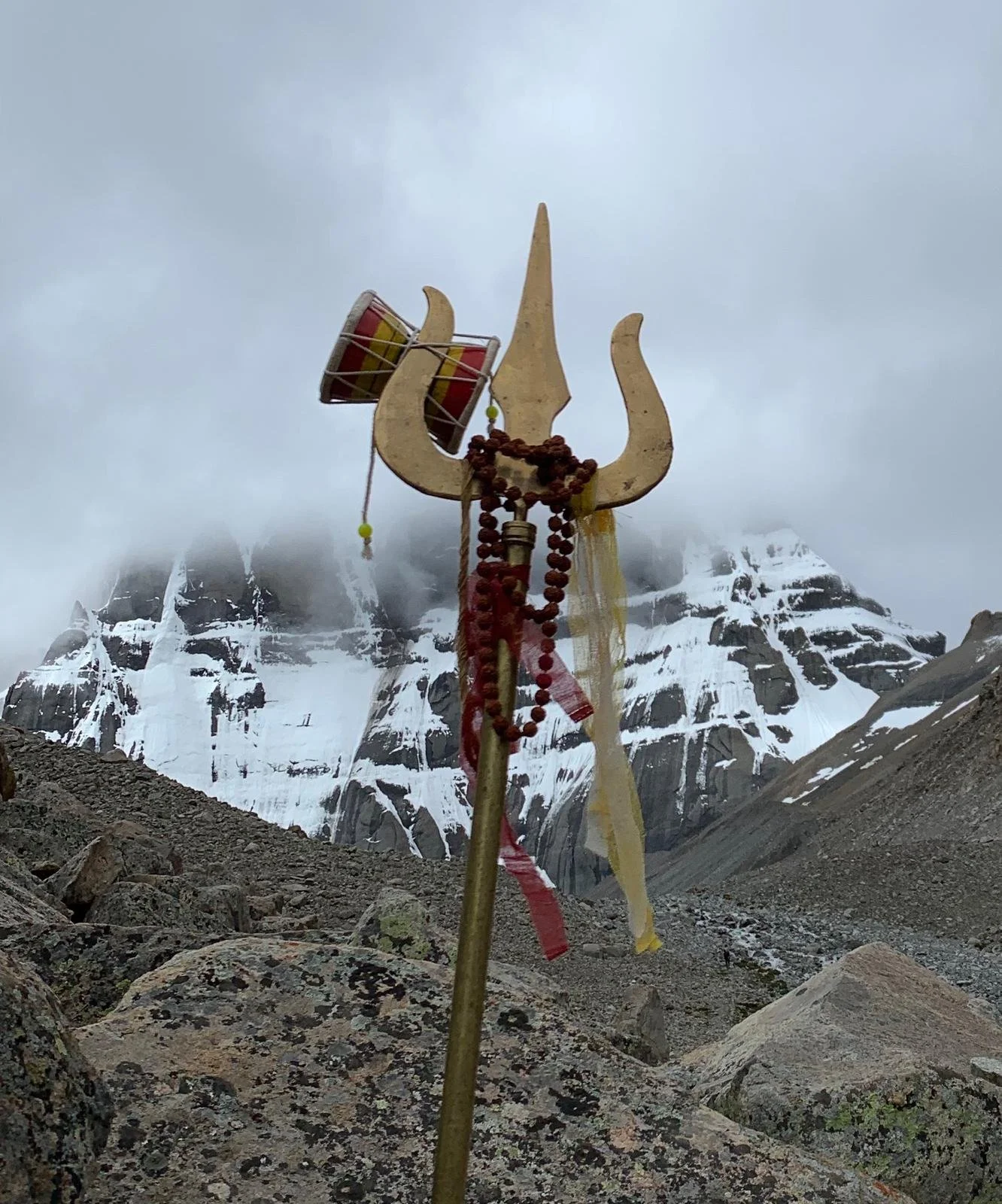 Mount Kailash with mist and snow-capped peaks, prayer flags, and a trident-shaped wooden structure with prayer beads and fabric strips.