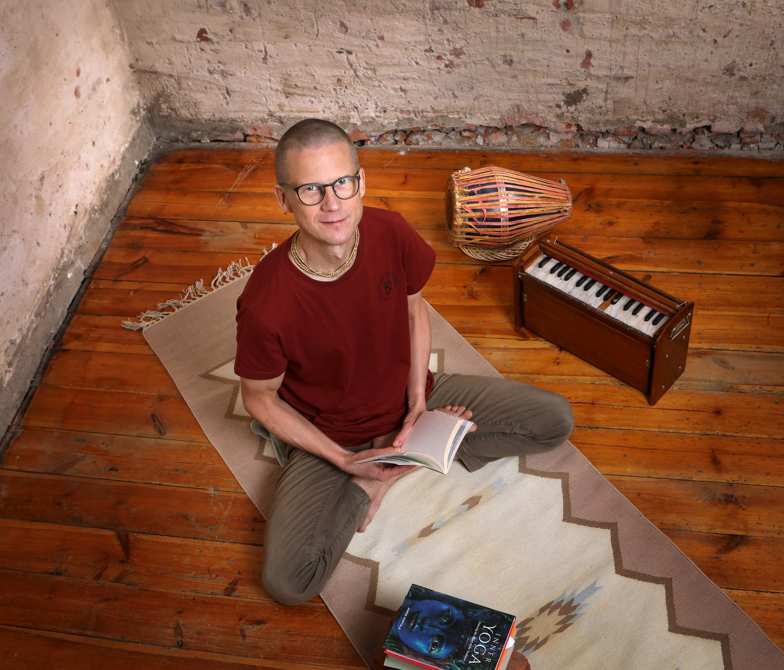A man with glasses, short hair, wearing a red t-shirt with a beaded necklace, is sitting on a beige rug on a wooden floor, holding a book open in his lap. Beside him is a small keyboard and a colorful drum. The background features an unfinished brick wall.