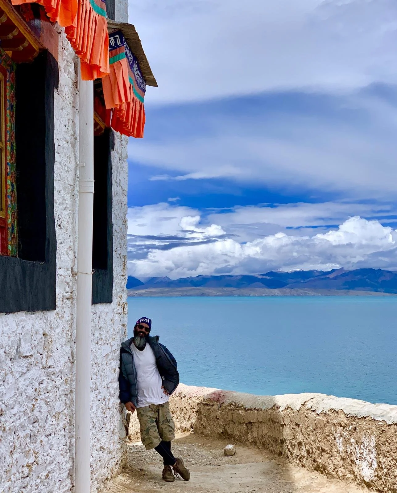 A man with a beard, wearing sunglasses, a cap, a gray jacket, and camouflage shorts, leaning against a stone building with a view of lake Monasarovar and mountains in the background. The building has colorful textiles hanging from the roof.