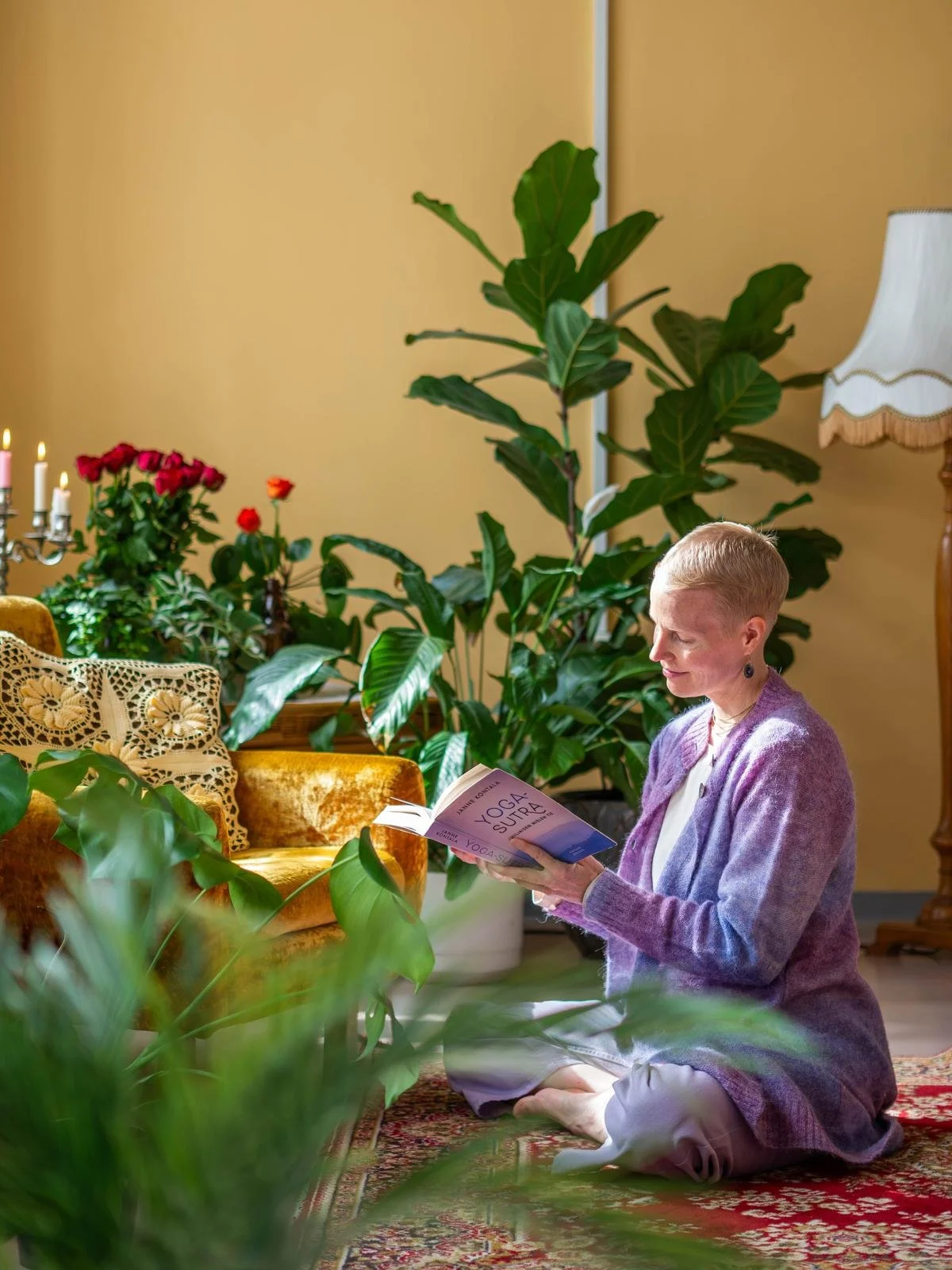 A woman sitting cross-legged on the floor reading a book titled 'Yoga Supta' in a cozy living room filled with green plants, a gold velvet sofa, and a lace pillow, with a yellow wall and a floor lamp in the background.