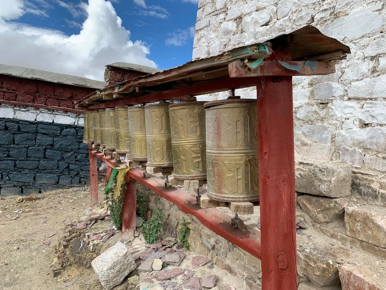 A row of prayer wheels mounted on a red wooden frame against a stone wall, with some greenery and rocks in the foreground.