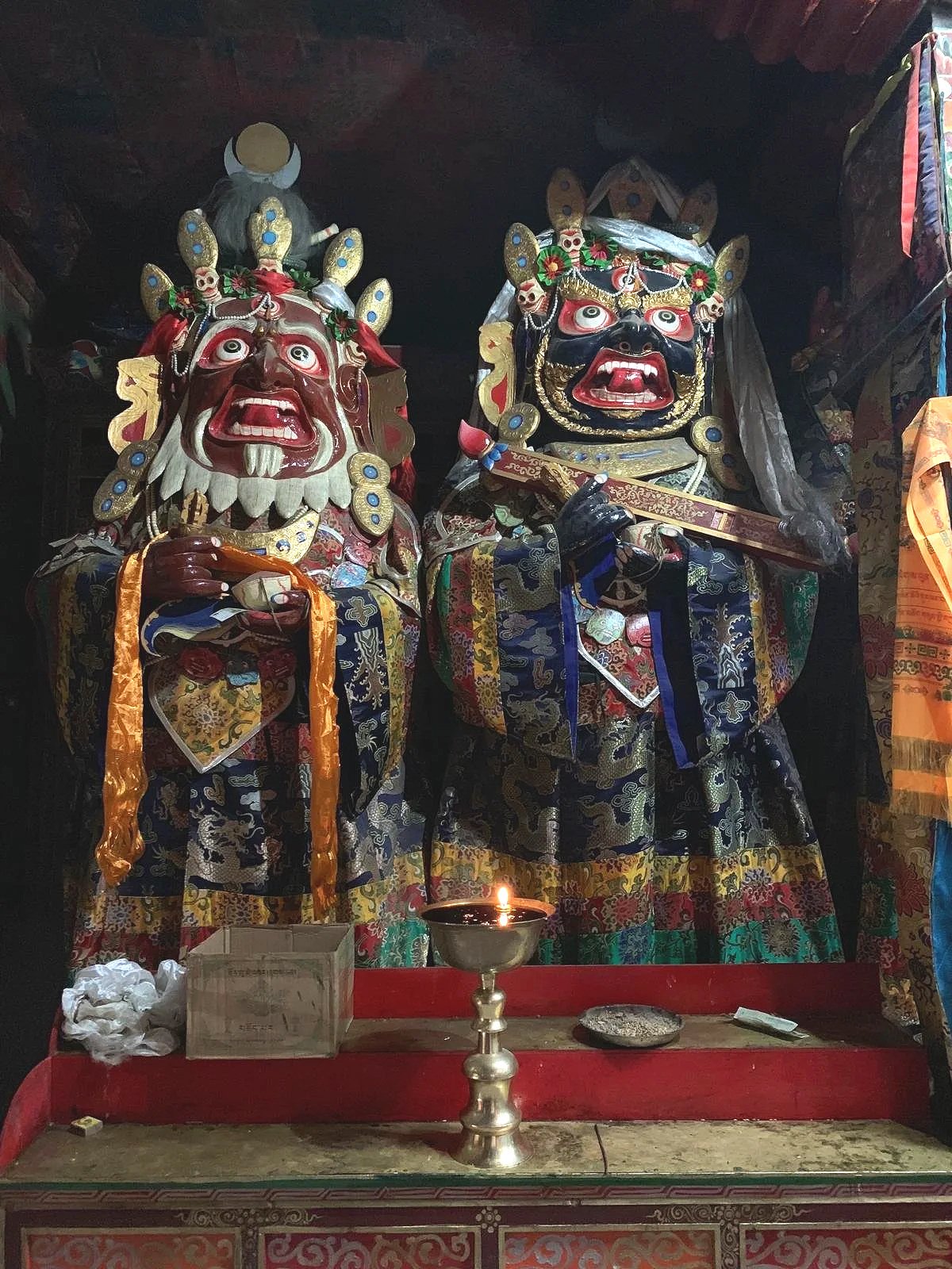 Two colorful traditional masks with fierce expressions and elaborate headpieces, dressed in ornate clothing, standing on an altar with a lit candle, offering box, and other ritual items.