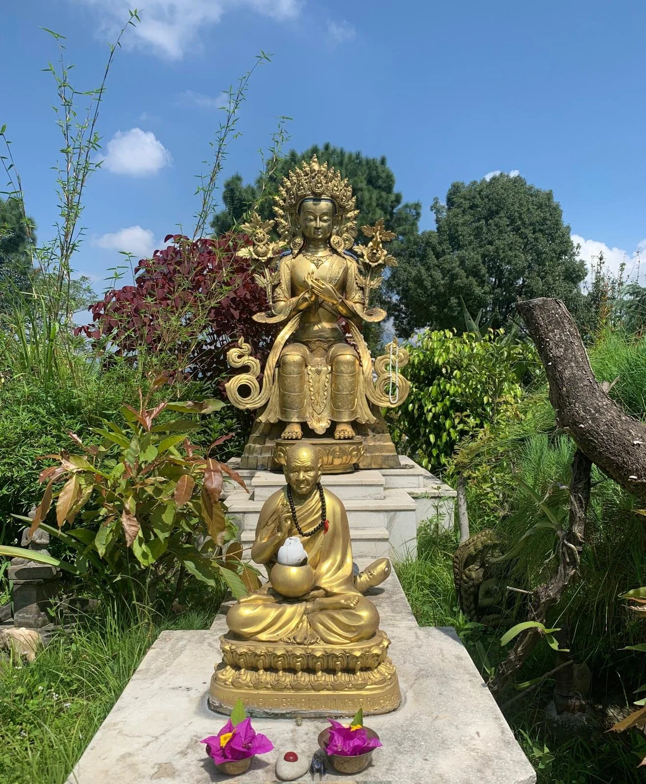 Golden statues of Buddhist deities and monks outdoors surrounded by lush greenery under a blue sky, with pink flowers and small offerings at the base.