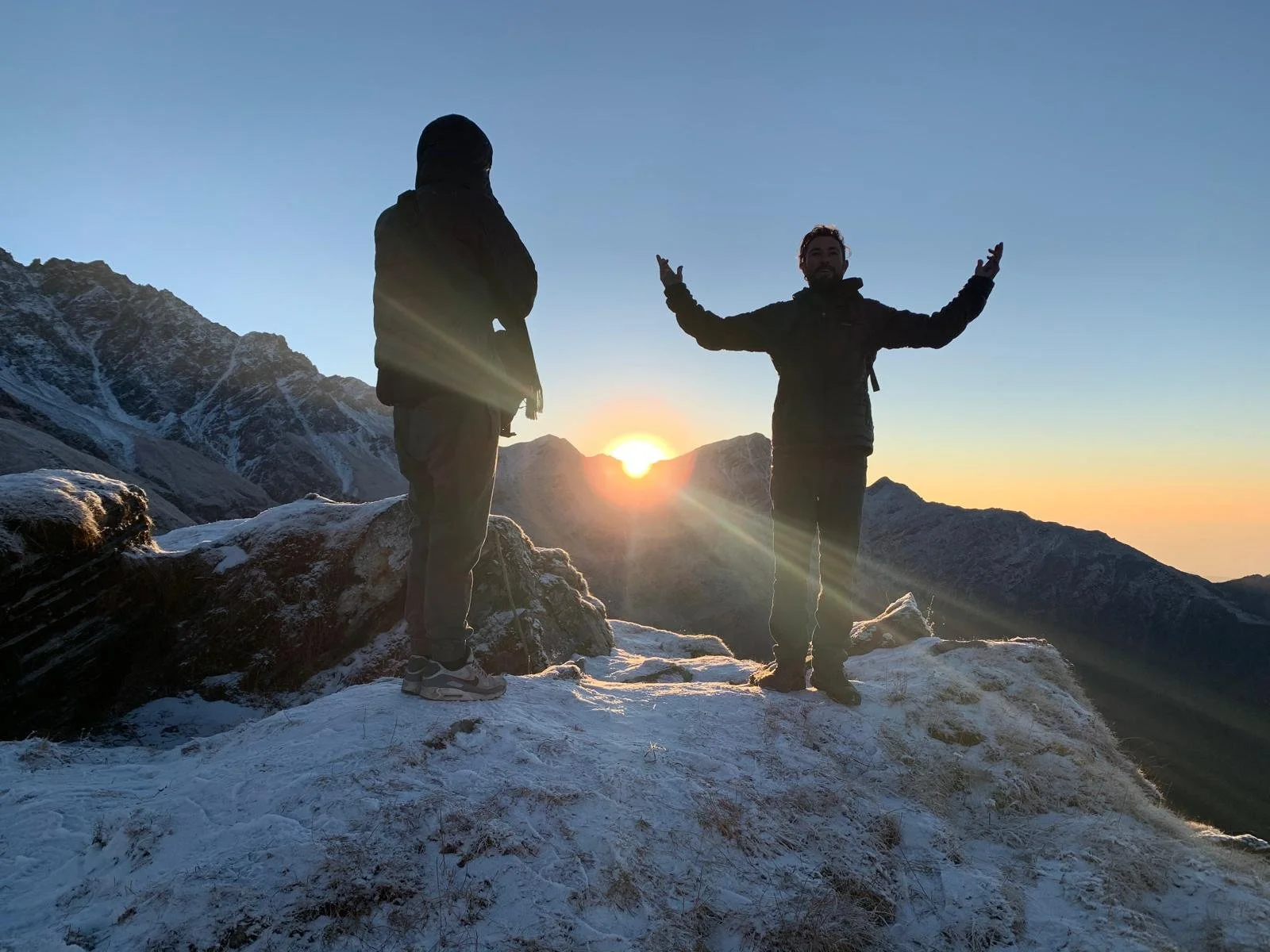 Two hikers standing on snowy mountain peak at sunrise, with one gesturing outward and the other facing away, with snow-covered mountains in the background.