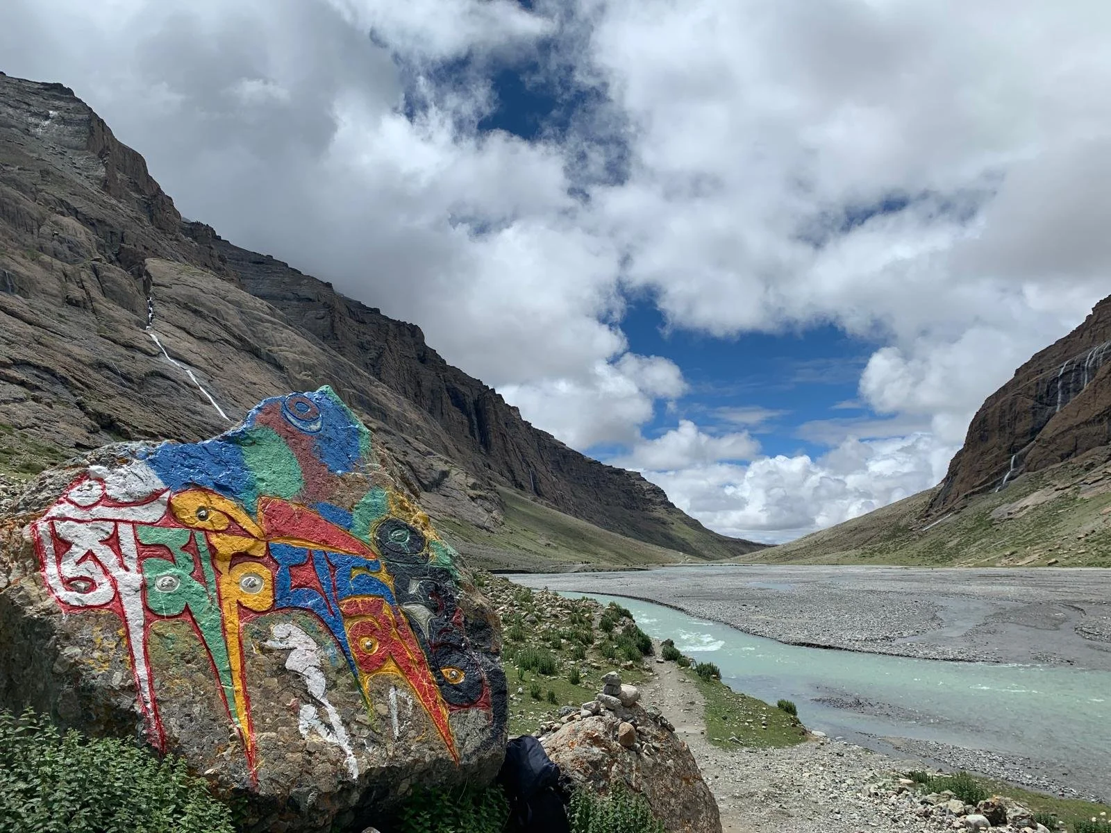 Colorfully painted rock with elephant design and script in front of a river valley with mountains and waterfalls under a partly cloudy sky.