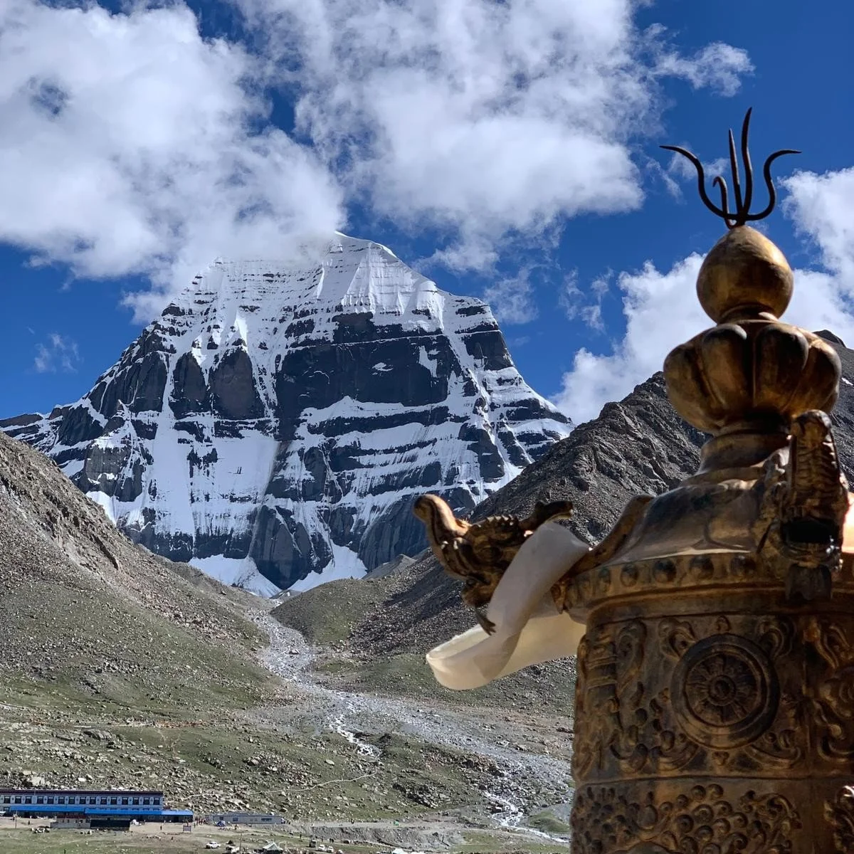 Snow-capped mountain with rugged slopes, partly cloudy sky, and a decorative golden religious statue in the foreground.