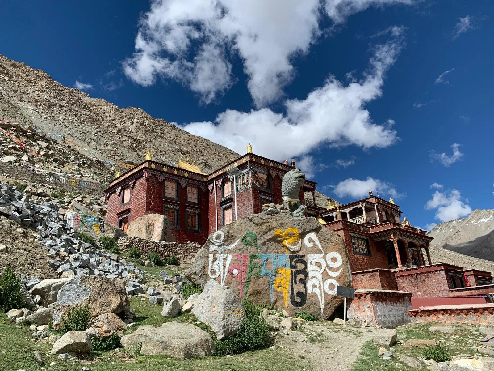 A Buddhist monastery built into a rocky mountain slope under a bright blue sky with clouds. The structure is red with gold accents and multiple levels with intricate window frames. In front, a large stone with colorful Tibetan symbols and a carved animal head sculpture is visible, surrounded by rocks and sparse greenery.