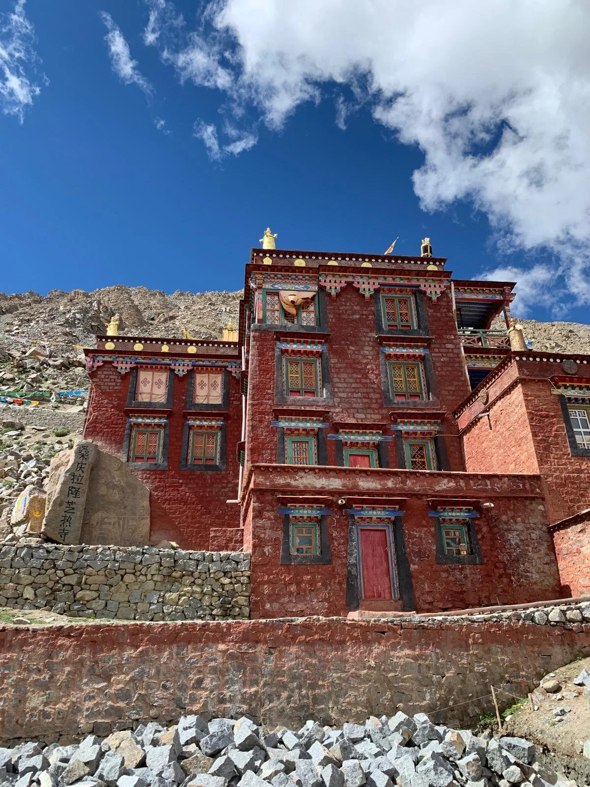 A multi-story red building with ornate, colorful window frames and decorations, situated on a rocky hillside under a blue sky with clouds.