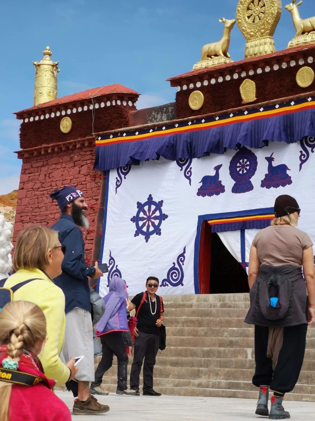 People standing in front of a structure with colorful decorations, including a large banner with traditional symbols and golden statues on top, against a clear blue sky.
