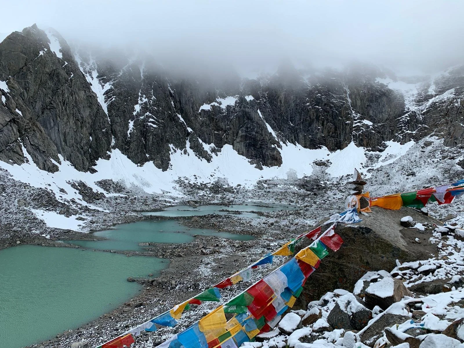 Mountain landscape with snow-covered rocks, turquoise lakes, and foggy peaks, decorated with colorful prayer flags.