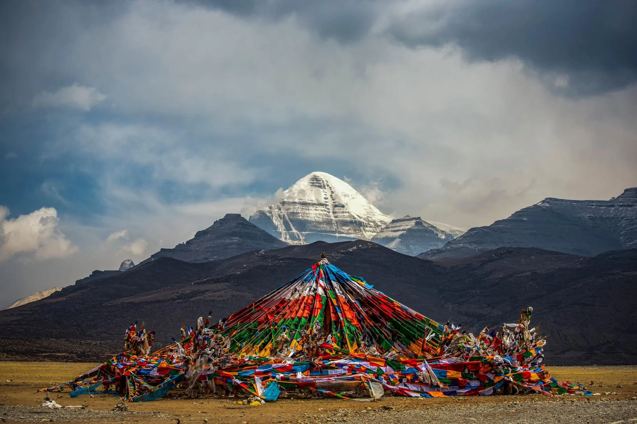 Colorful prayer flags draped over a structure with snow-capped mountain in the background, possibly Mount Everest, under a cloudy sky.