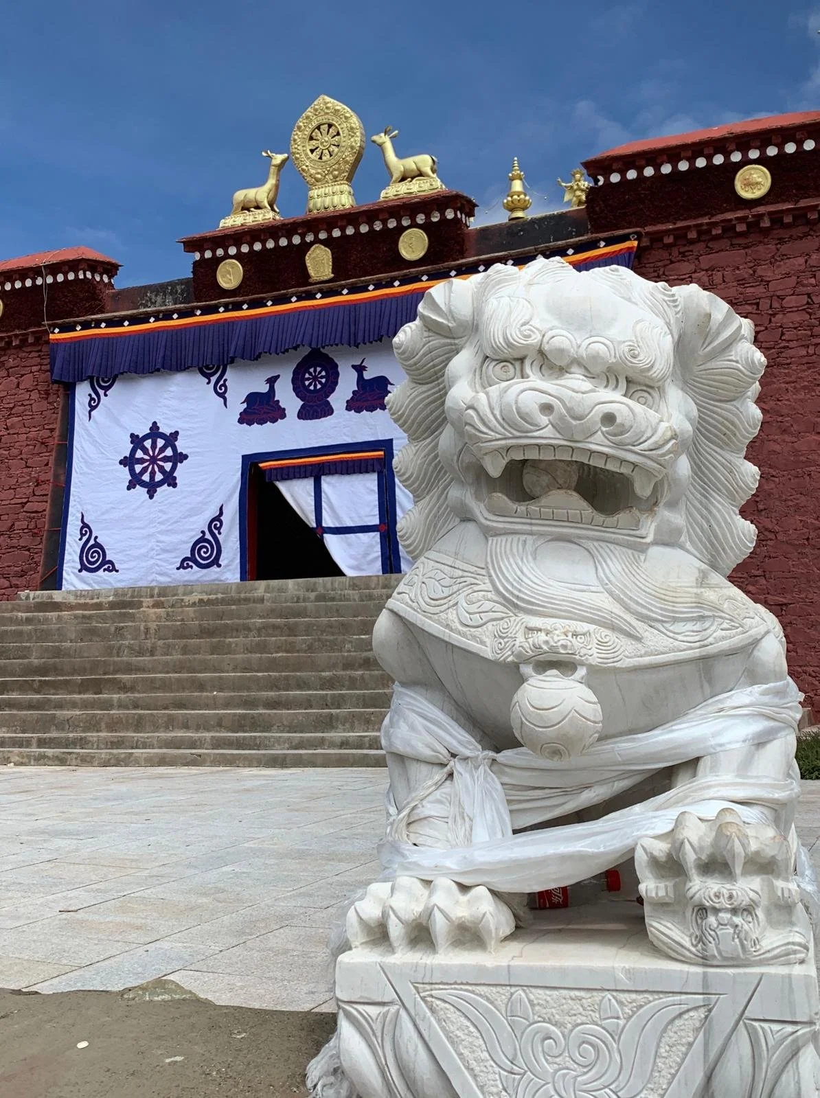 White stone lion statue in front of a building with steps, decorated with traditional Tibetan symbols and gold ornaments, featuring a blue and white tapestry above the entrance.