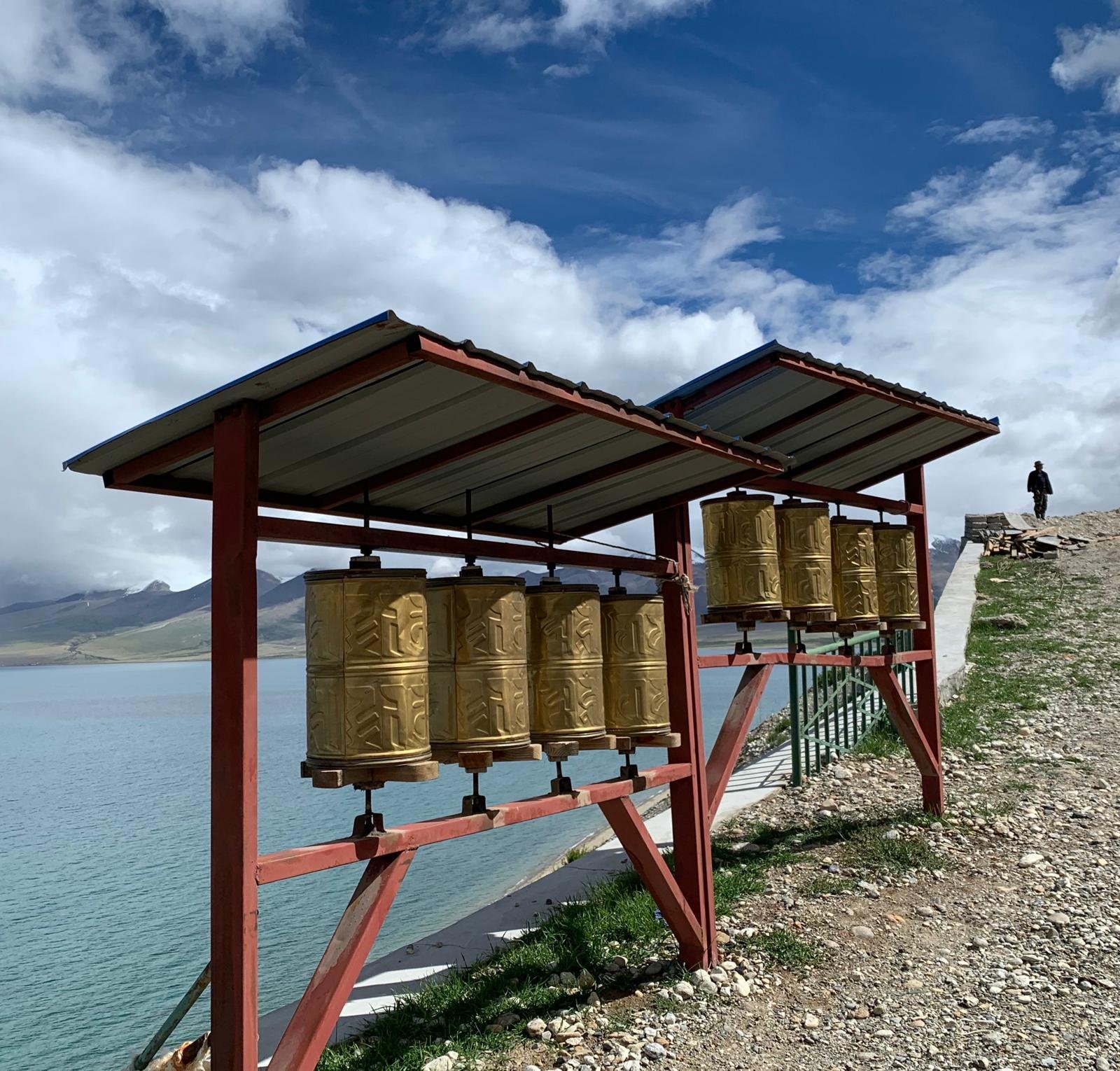 A row of large prayer drums with symbols, under a metal roof on a red wooden structure, overlooking a body of water with mountains in the distance, and a person walking along the hill.