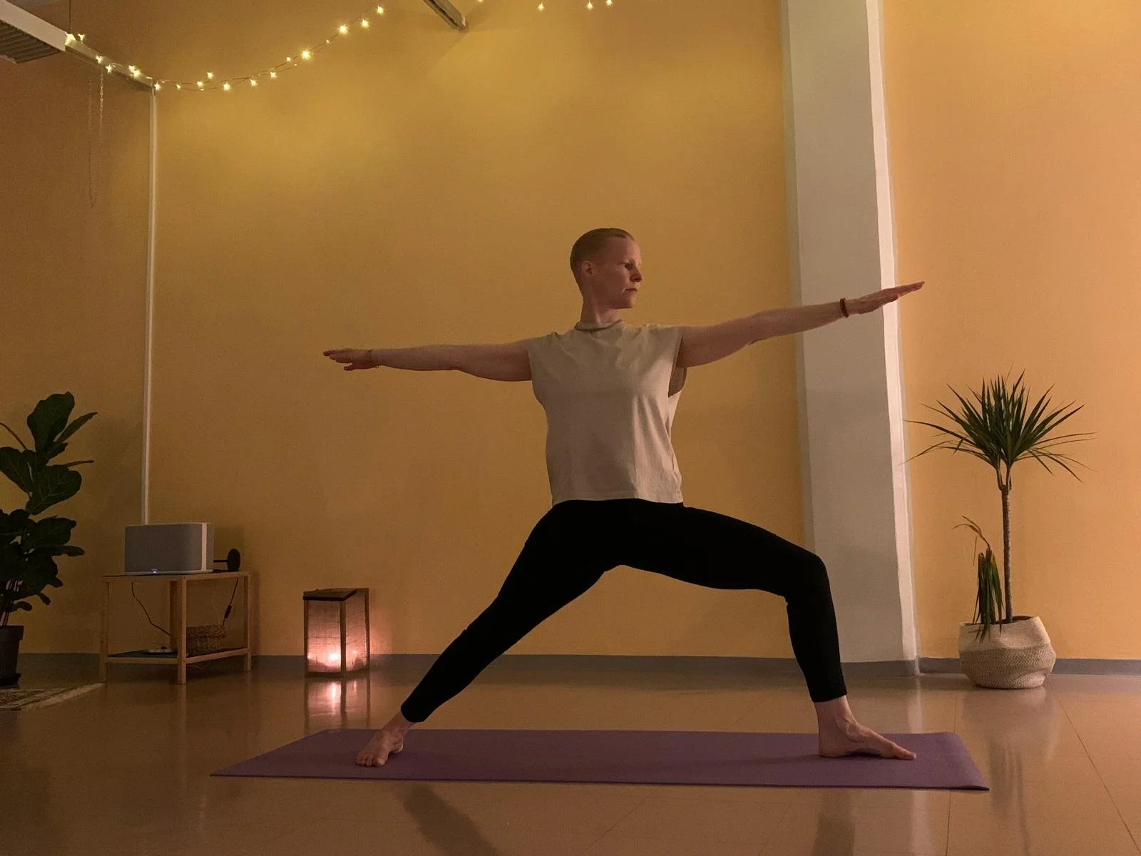 A woman practicing yoga in a dimly lit room decorated with potted plants, string lights, and candles. She is standing on a yoga mat, in a warrior pose with arms extended and legs bent.