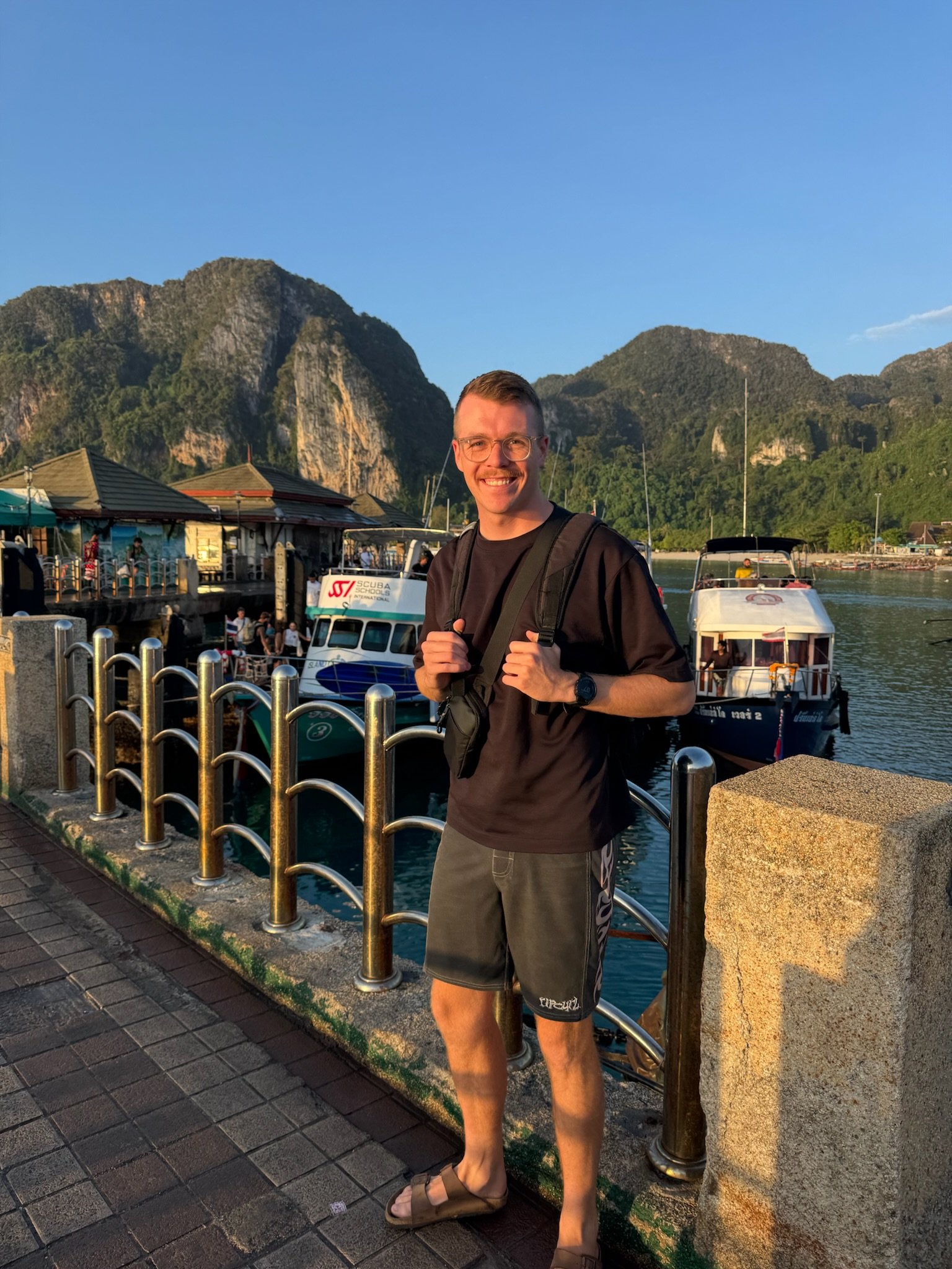 A man smiling with a backpack standing on a waterfront promenade with boats and a mountain in the background during sunset.
