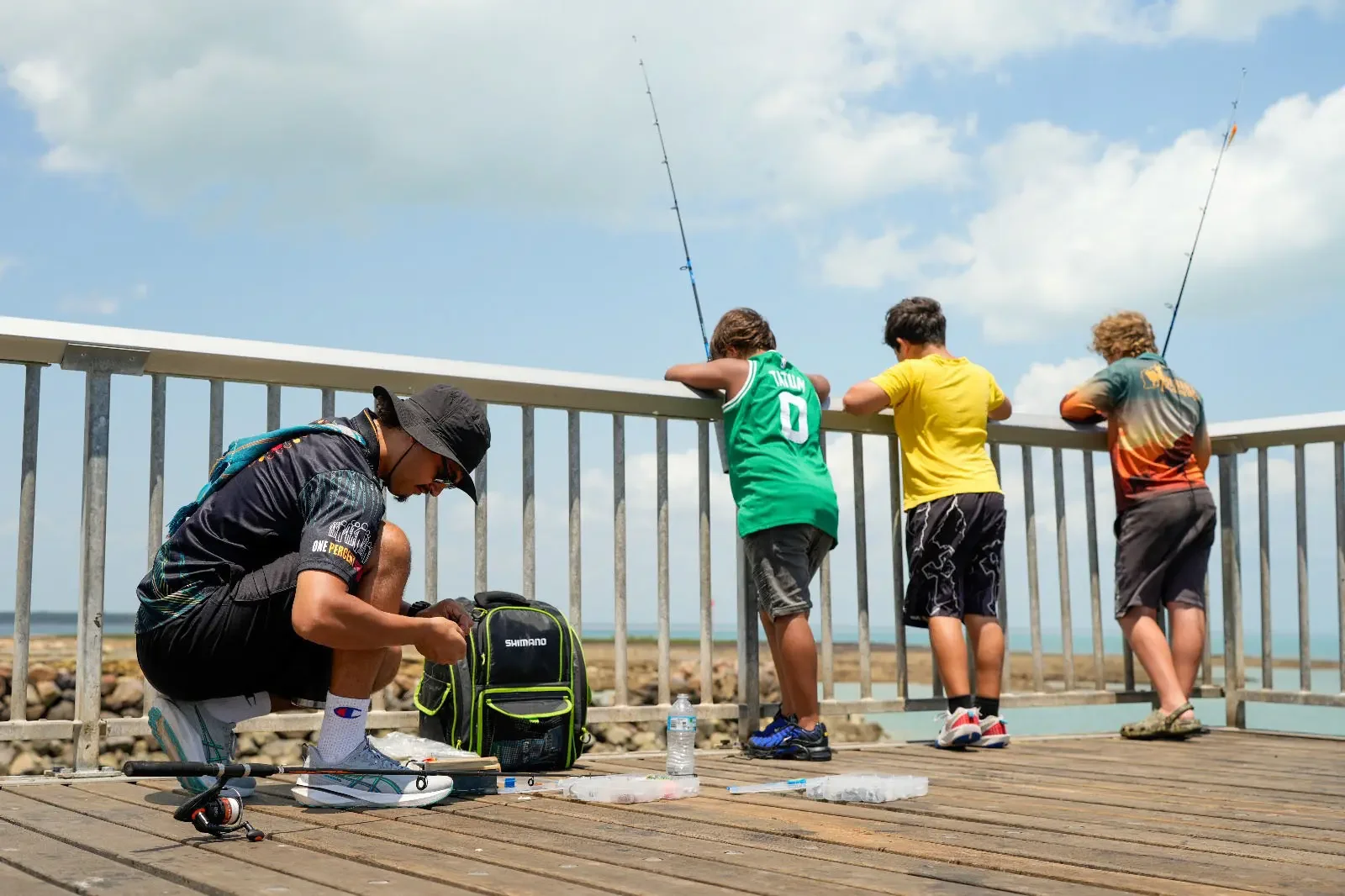 A man crouching on a wooden dock near the water, working on fishing gear with a Shimano backpack nearby. Three boys stand at the edge of the dock, looking out at the water with fishing rods in hand, under a partly cloudy sky.