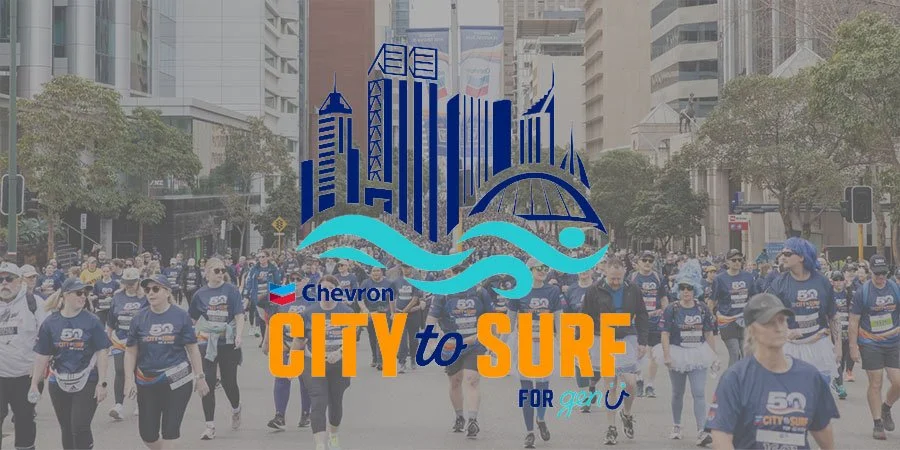 People participating in a City to Surf race on a city street, with a city skyline and trees in the background. The event is sponsored by Chevron.
