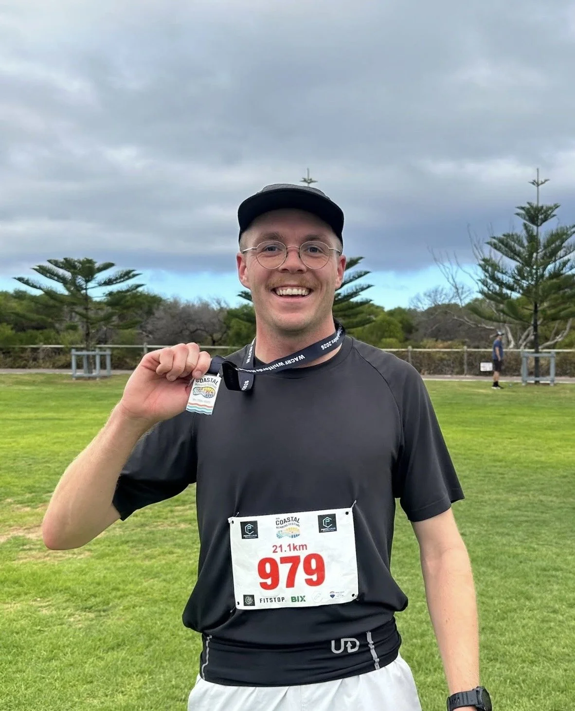 Man holding race medal, standing on grassy field with trees, cloudy sky in background, wearing black athletic clothes with race bib 979, 21.1 km.