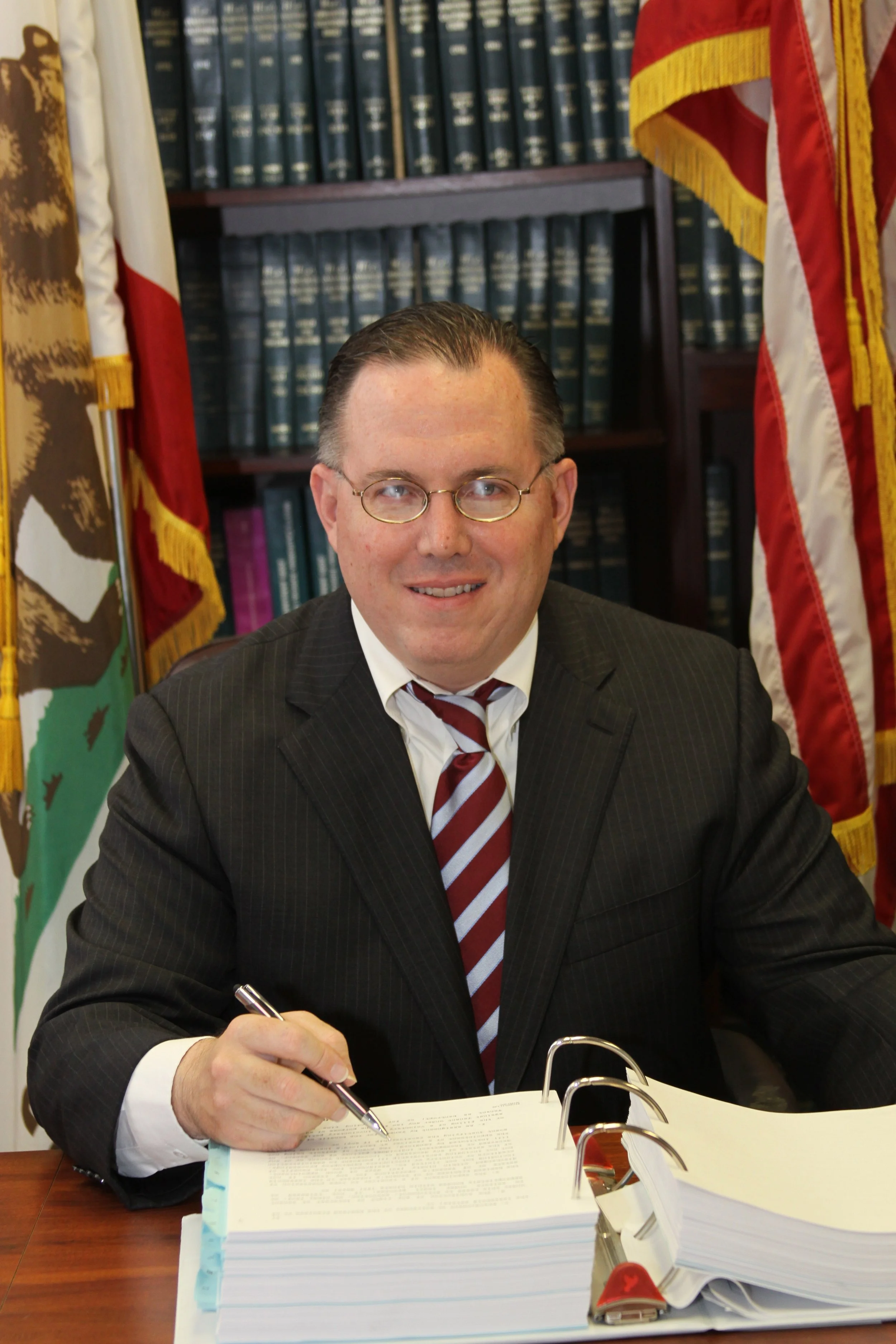Charles E. Pell seated at a desk with law books and flags, candidate for Orange County Superior Court Judge.