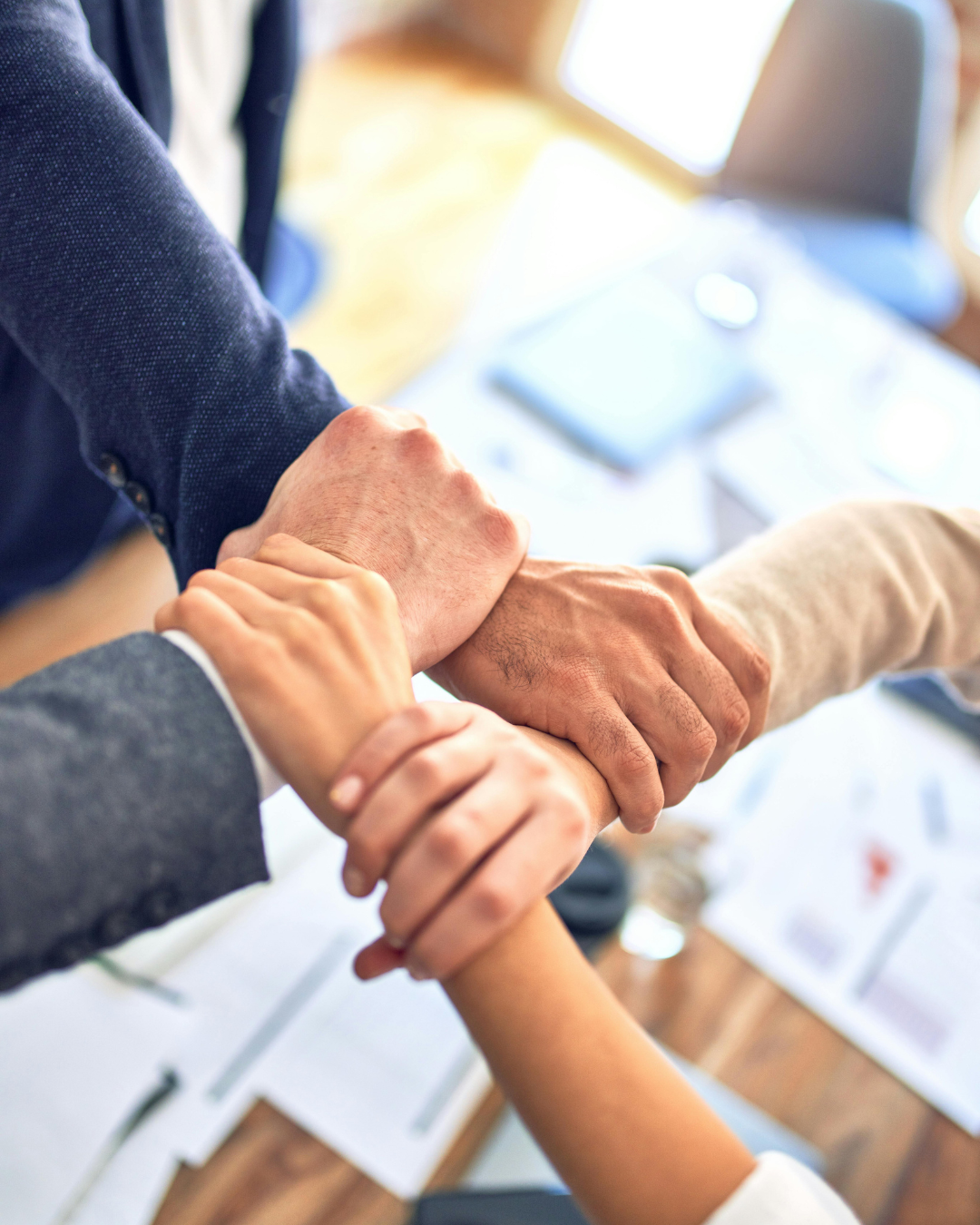 Multiple hands joined in a handshake over a desk with papers and a laptop.