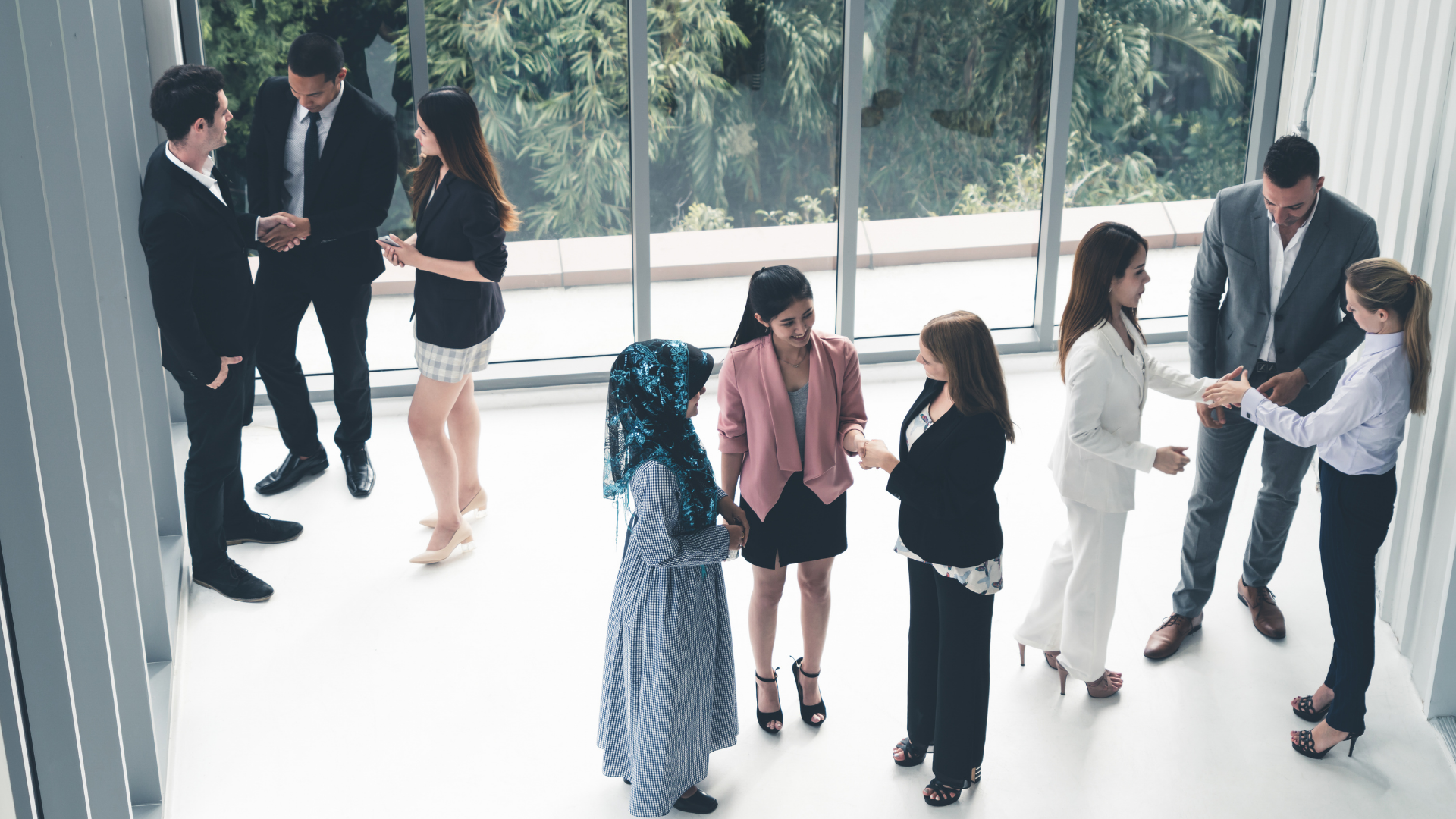 Group of diverse professionals at a business networking event, greeting each other with handshakes and conversations indoors near large windows.