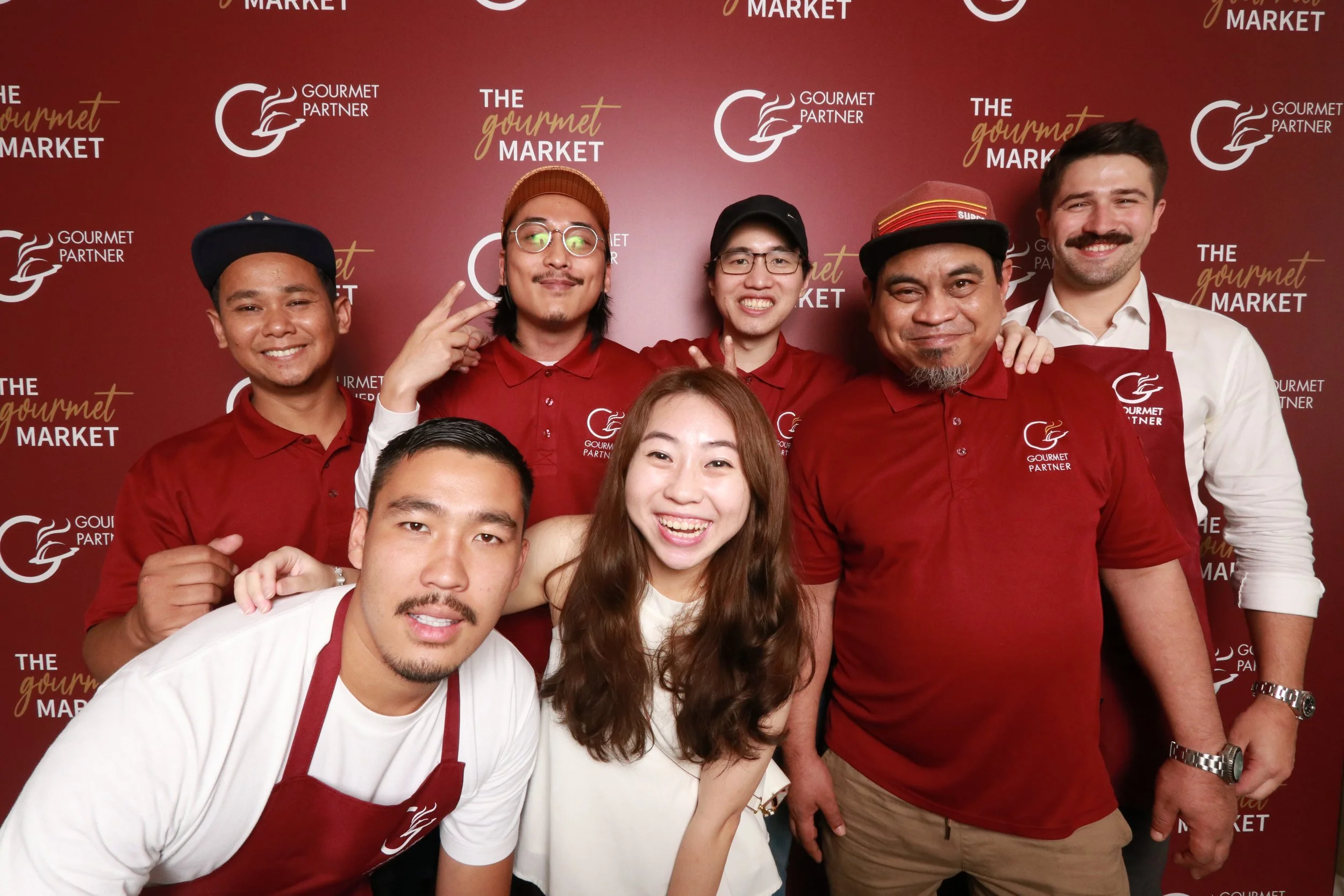 Group of six smiling people standing in front of a red backdrop with logos and text, some wearing red shirts and aprons, posing for a photo at a gourmet market event.