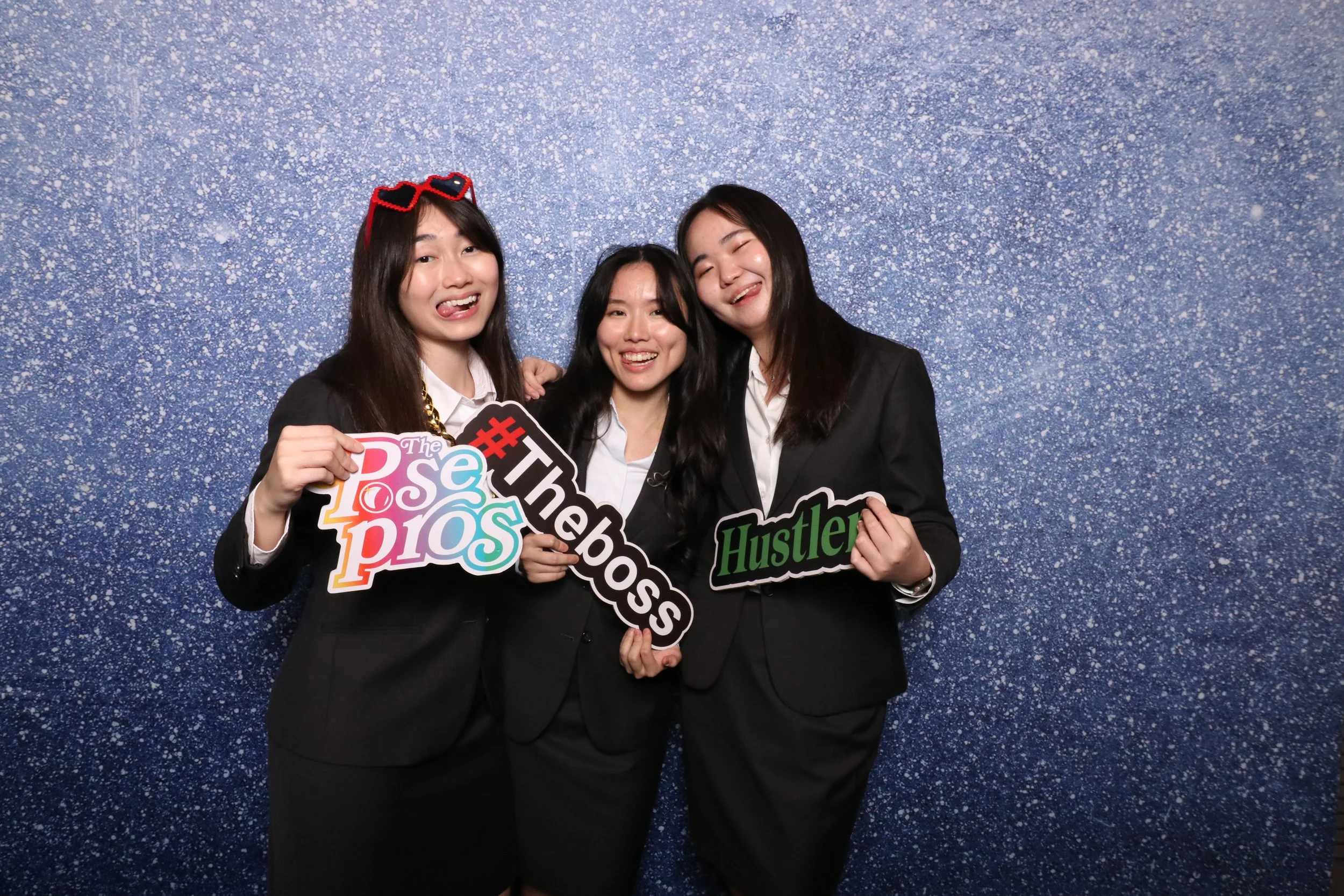 Three young women in business suits standing together in front of a blue, sparkly backdrop at an event, holding colorful signs with words and hashtags, smiling and posing for the camera.